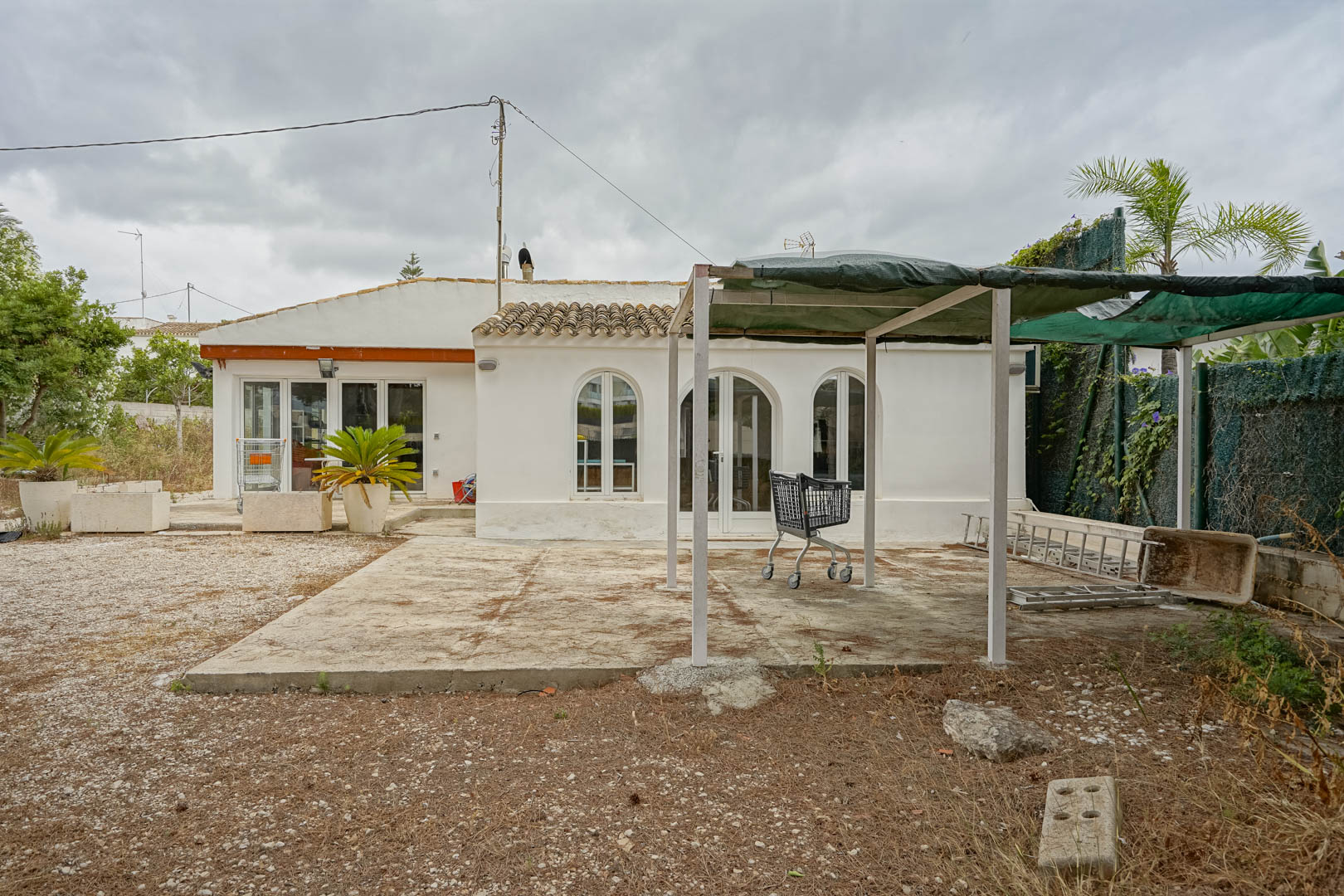 Vue extérieure de la villa de plain-pied avec fenêtres cintrées, cour en gravier et structure de pergola métallique avec toile d'ombrage.