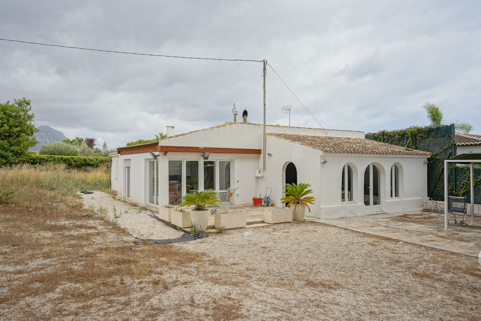 Vue extérieure de la villa méditerranéenne avec arches traditionnelles, menuiseries blanches et toit en tuiles, sur un vaste terrain plat avec vue montagne.