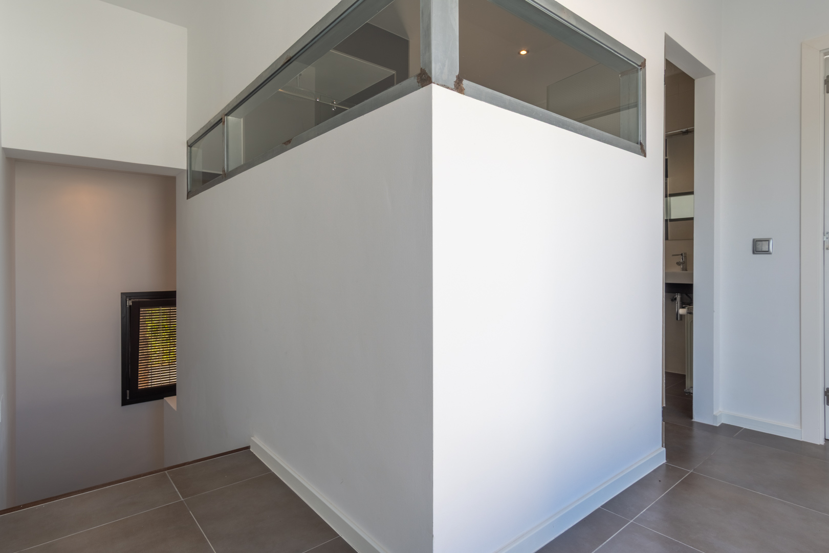 Couloir intérieur avec carrelage gris, cloison avec imposte vitrée en métal et accès à l'escalier menant au niveau inférieur et à une salle d'eau.