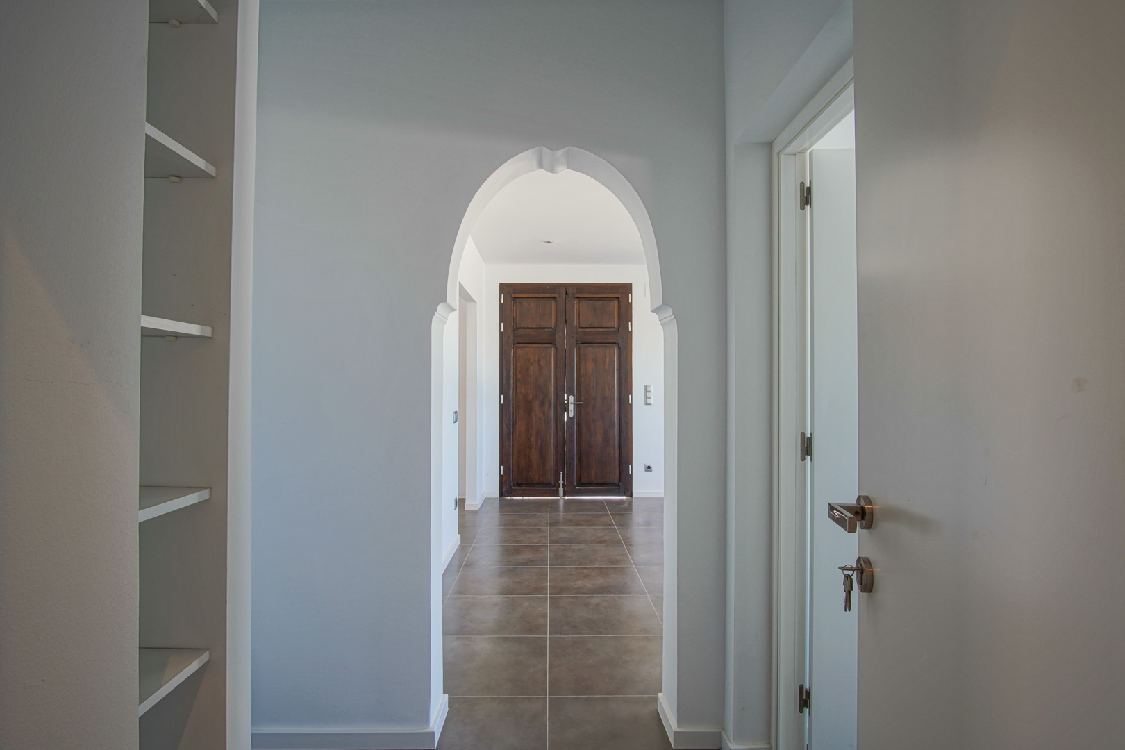 Couloir intérieur avec une arche de style méditerranéen encadrant une double porte en bois sombre restaurée sur un sol en grès cérame.