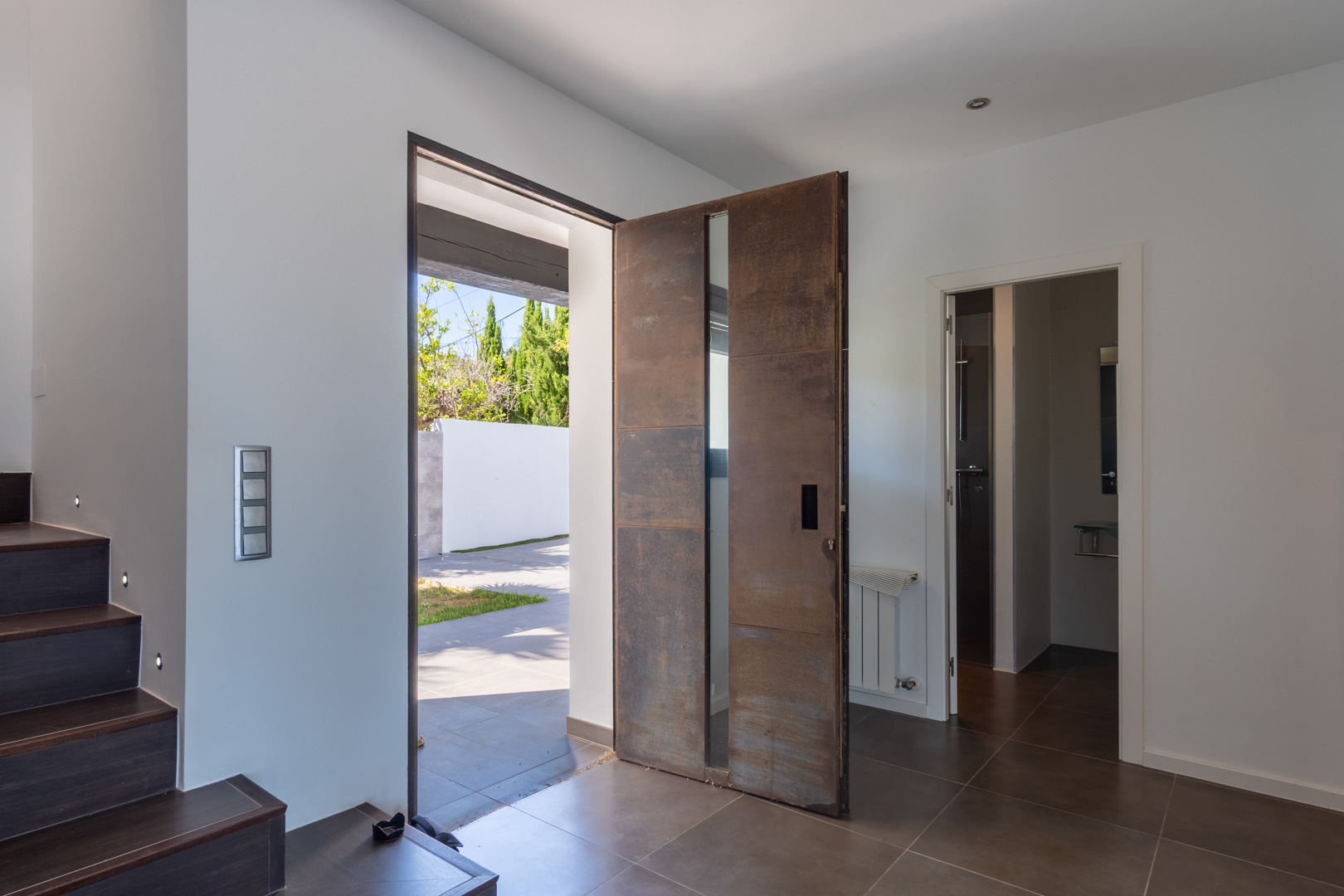 Entrée principale avec porte pivotante en acier corten et insert vitré. L'espace propose un sol sombre, un escalier avec balisage LED et un accès à une salle d'eau.