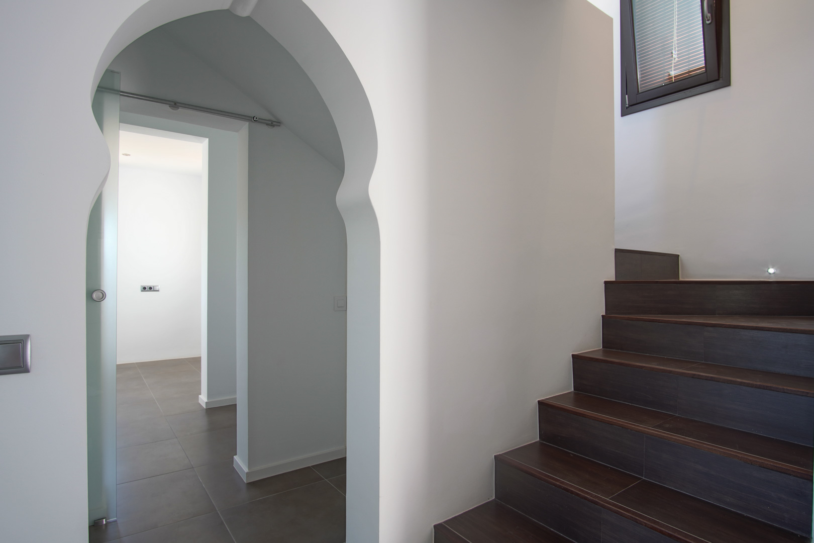 Escalier intérieur avec marches en grès foncé et éclairage intégré, à côté d'une arche de style mauresque menant à une pièce avec porte coulissante en verre.