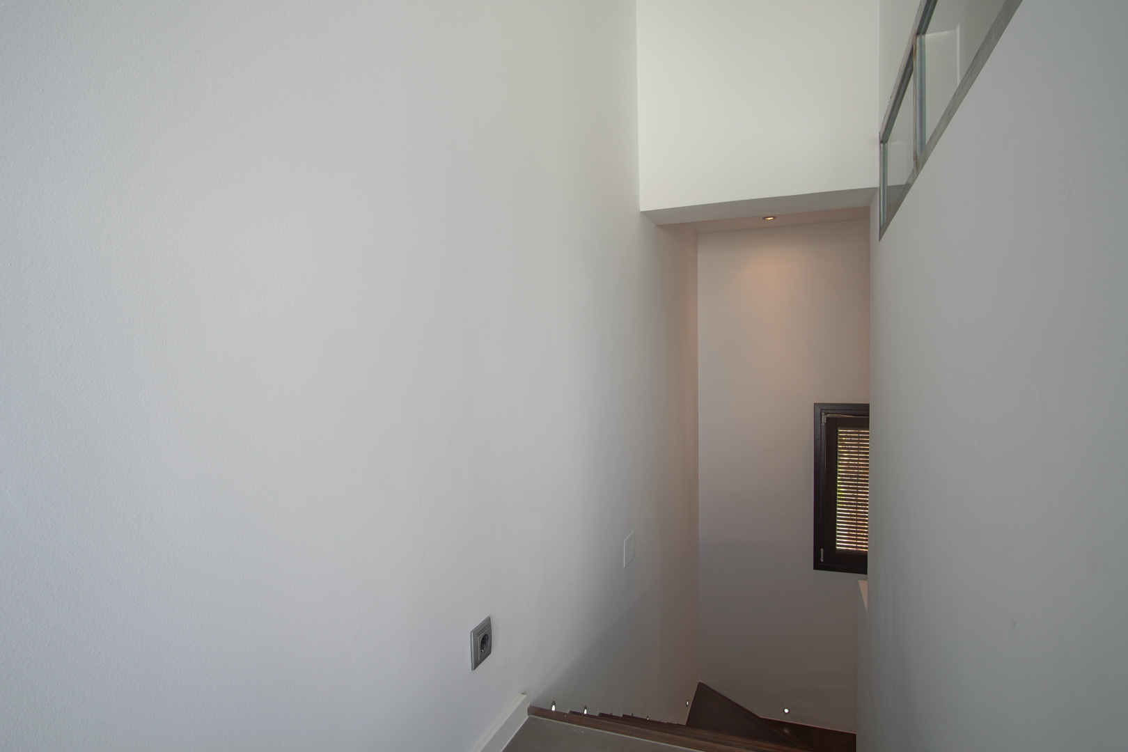 Escalier intérieur avec marches sombres et éclairage LED intégré au sol. Murs blancs épurés et petite fenêtre avec cadre sombre et volets à persiennes.