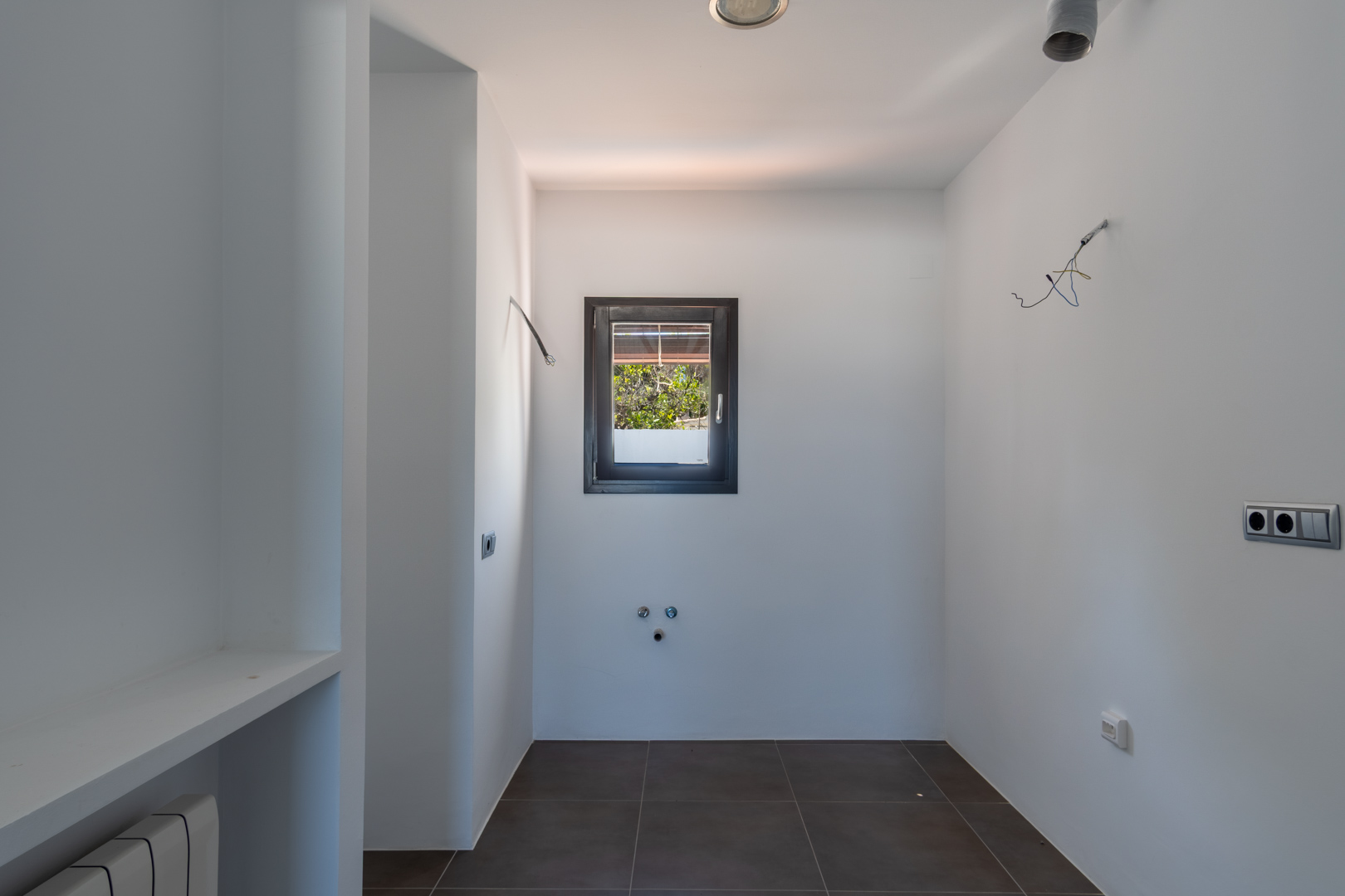 Espace buanderie avec pré-installations de plomberie et d'électricité, carrelage au sol foncé et fenêtre à cadre sombre ouvrant sur l'extérieur verdoyant.
