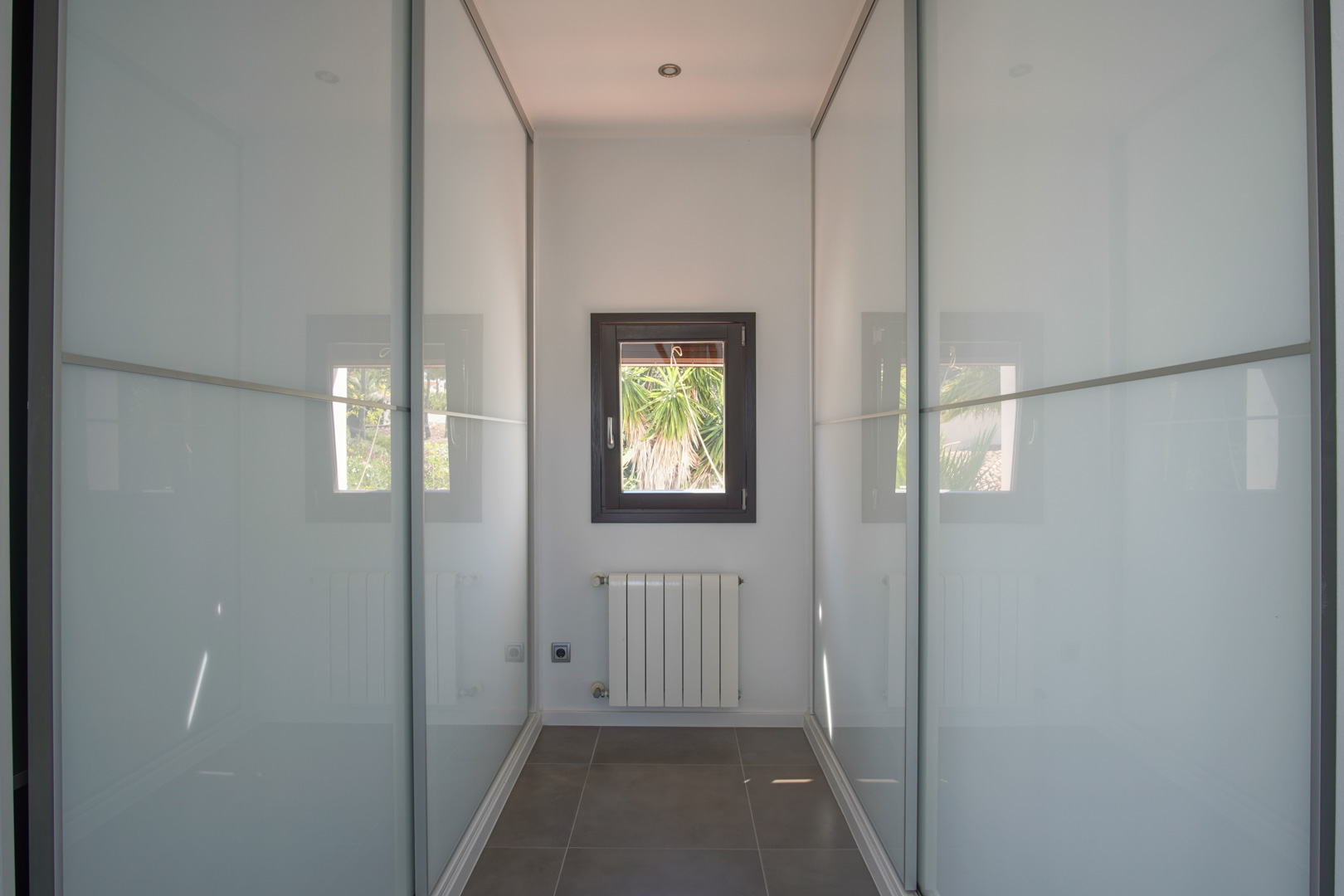 Espace dressing avec placards intégrés à portes coulissantes en verre dépoli, radiateur mural et fenêtre cadrant la végétation extérieure.