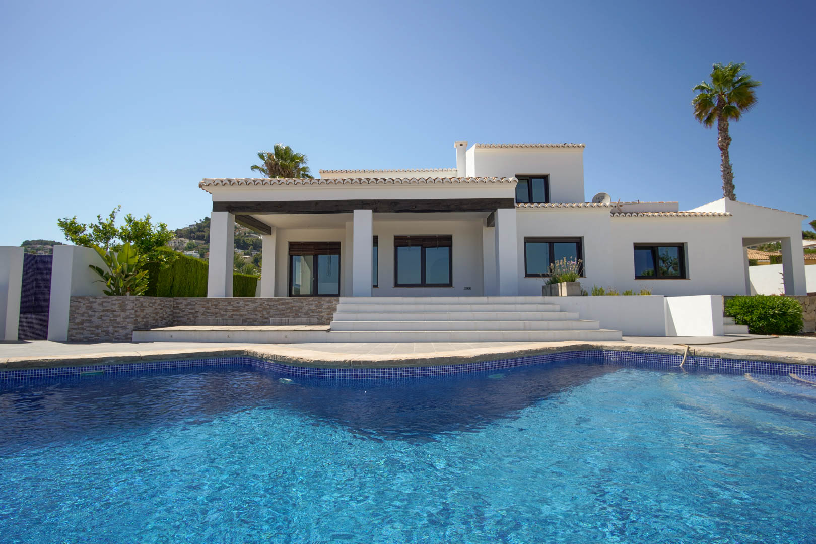 Façade de villa méditerranéenne blanche avec piscine en mosaïque bleue. Comprend un porche couvert aux poutres en bois sombre et de larges escaliers d'accès.