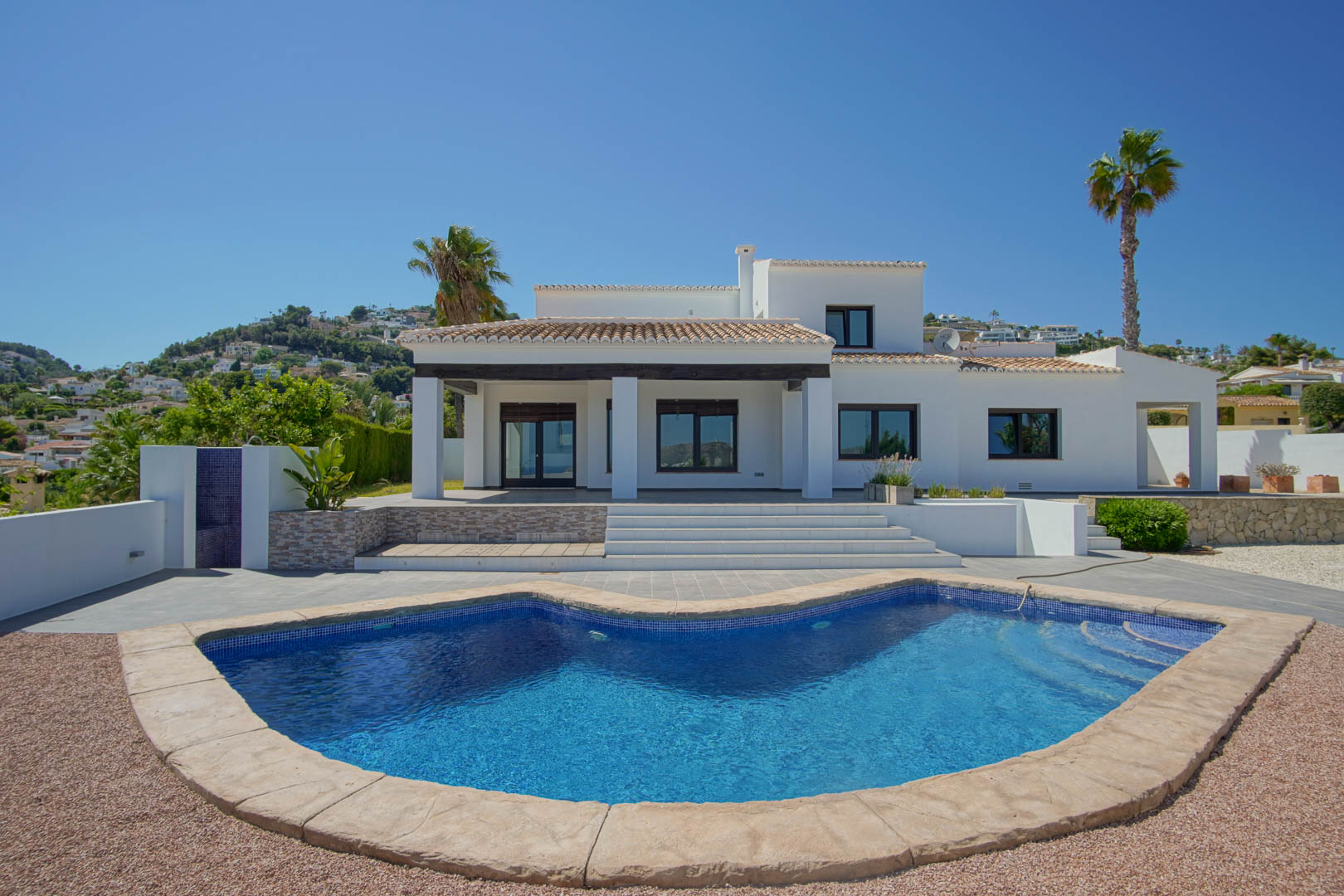 Façade principale de la villa avec piscine aux formes courbes, porche couvert à poutres en bois sombre et vastes terrasses ensoleillées sous un ciel dégagé.