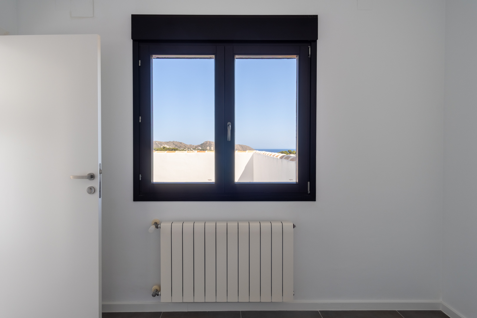 Fenêtre en aluminium sombre avec double vitrage offrant une vue sur la mer et le Cabo de Moraira, située au-dessus d'un radiateur blanc dans une pièce lumineuse épurée.