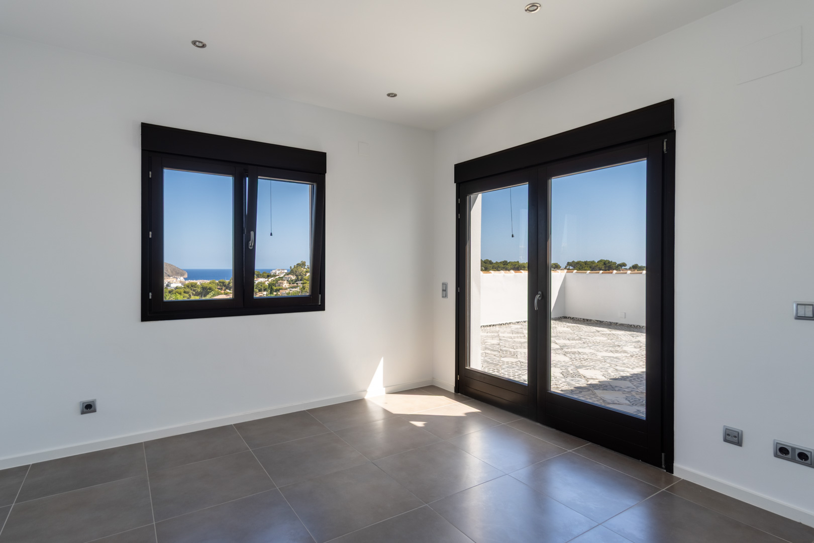 Pièce lumineuse avec carrelage gris, menuiseries sombres et accès direct à une terrasse offrant une vue panoramique sur la mer et le littoral.