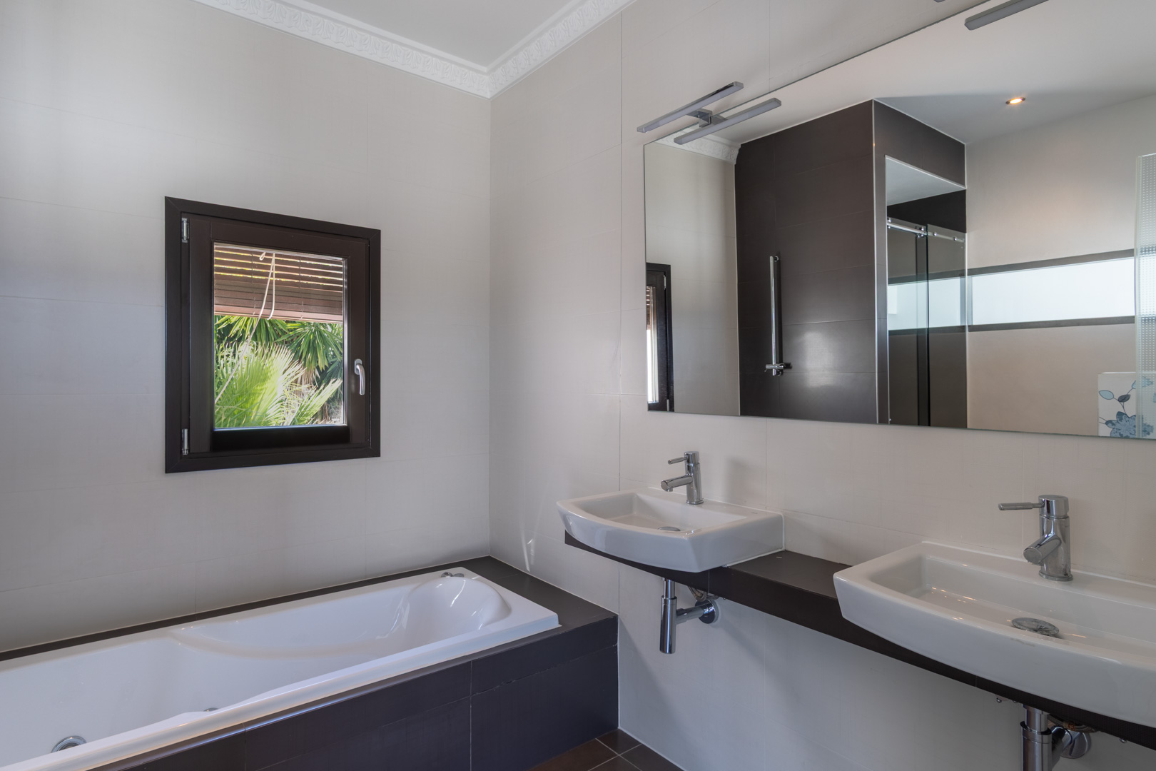 Salle de bains moderne avec double vasque sur plan sombre, baignoire intégrée et fenêtre à cadre effet bois offrant lumière naturelle et vue sur le jardin.
