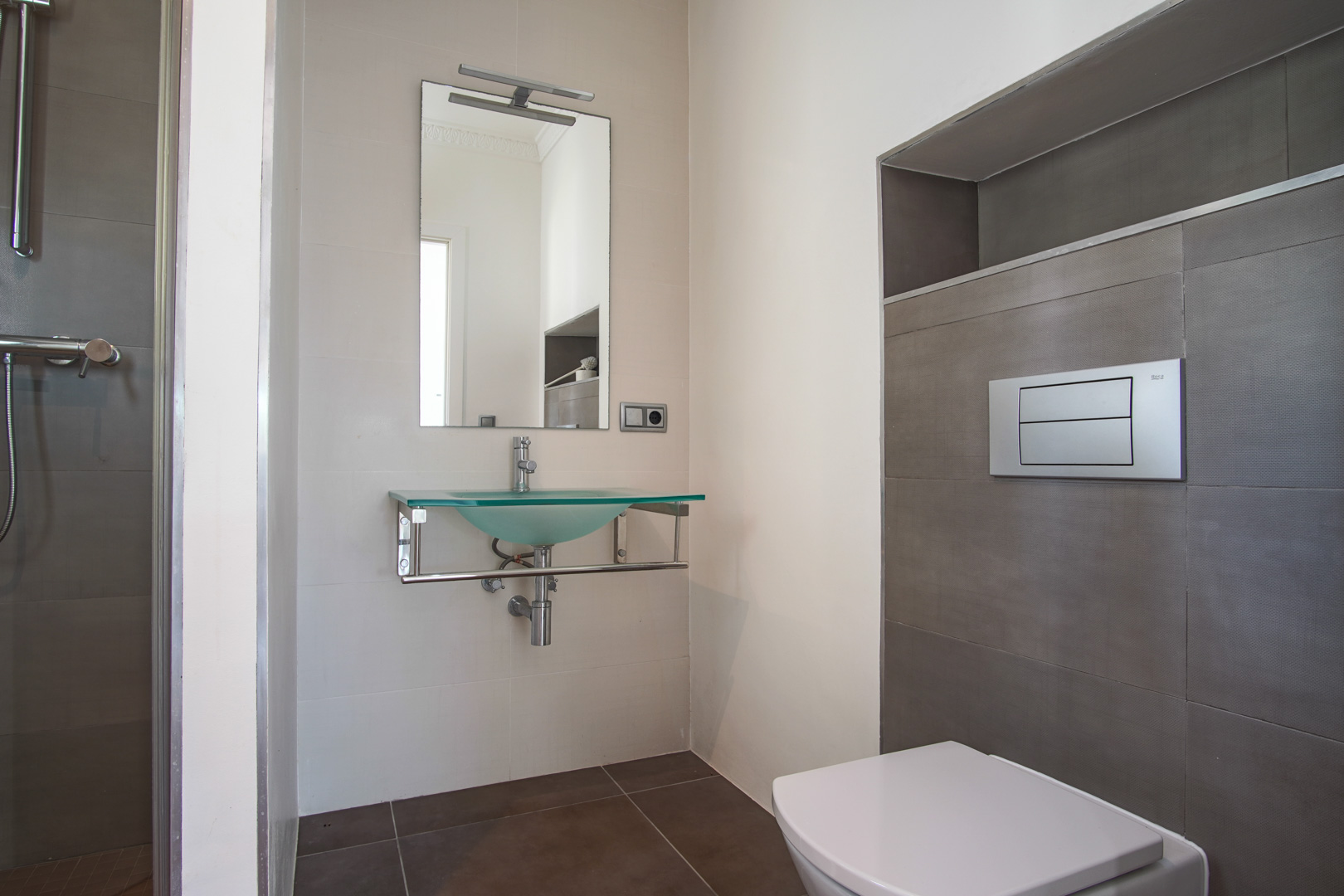 Salle de bains moderne avec WC suspendu, vasque en verre dépoli sur support chromé et carrelage texturé intégrant une niche de rangement murale.