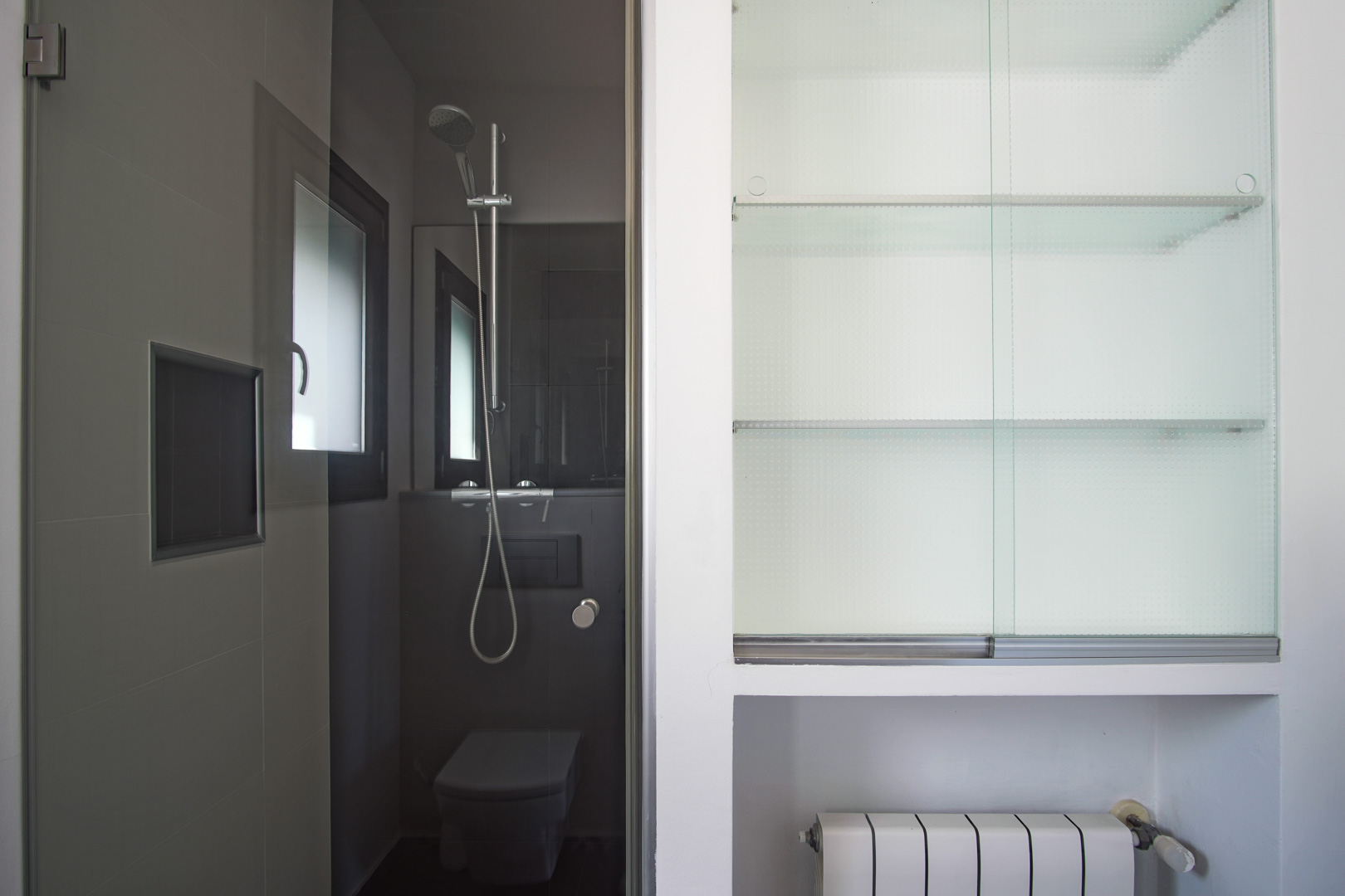 Salle d'eau moderne avec carrelage gris, douche à l'italienne et WC suspendu, complétée par un placard intégré à étagères en verre et un radiateur.