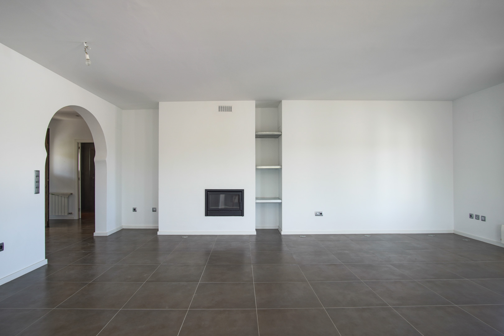 Salon ouvert avec cheminée encastrée, étagères intégrées et sol en grès cérame foncé. Une arche méditerranéenne relie l'espace au couloir et aux autres pièces.