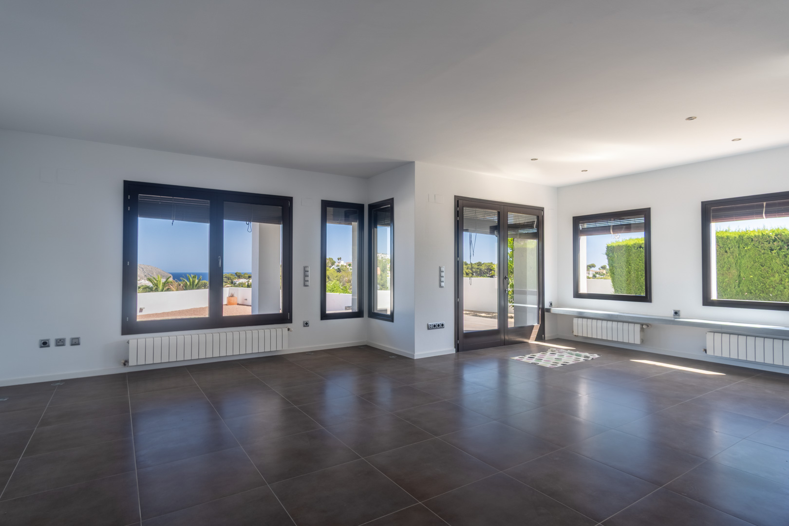 Vaste salon-salle à manger avec sol en grès cérame foncé, radiateurs et menuiseries en aluminium effet bois ouvrant sur des vues dégagées sur la mer.