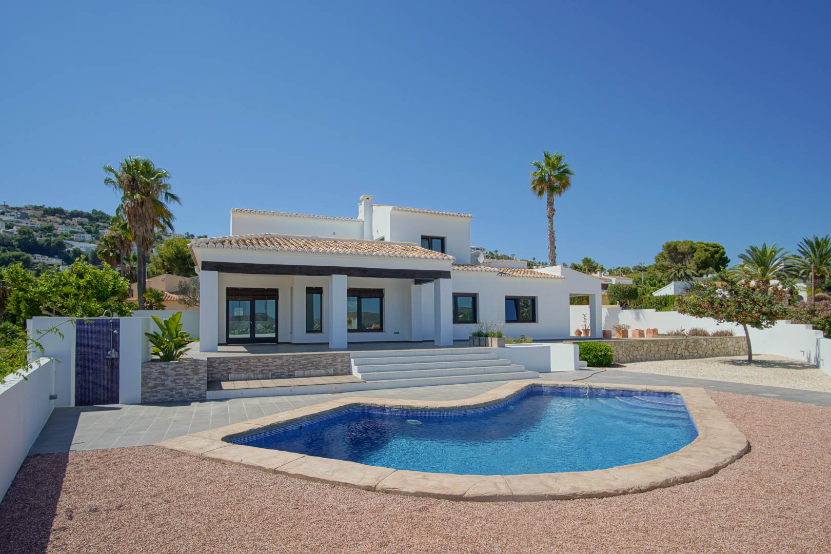 Vue extérieure de la villa avec sa piscine aux formes organiques, son jardin méditerranéen en gravier et sa terrasse couverte aux poutres en bois sombre.