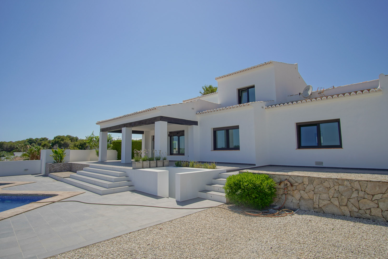 Vue extérieure de la villa méditerranéenne avec façade blanche, porche aux poutres sombres et terrasse en paliers menant à la piscine et aux murs en pierre.