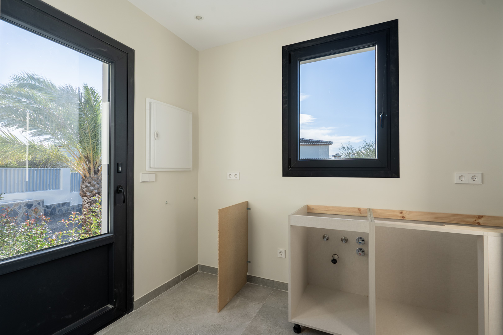 Détail de la buanderie avec porte sur le jardin, fenêtre à cadre noir et meuble bas en construction.