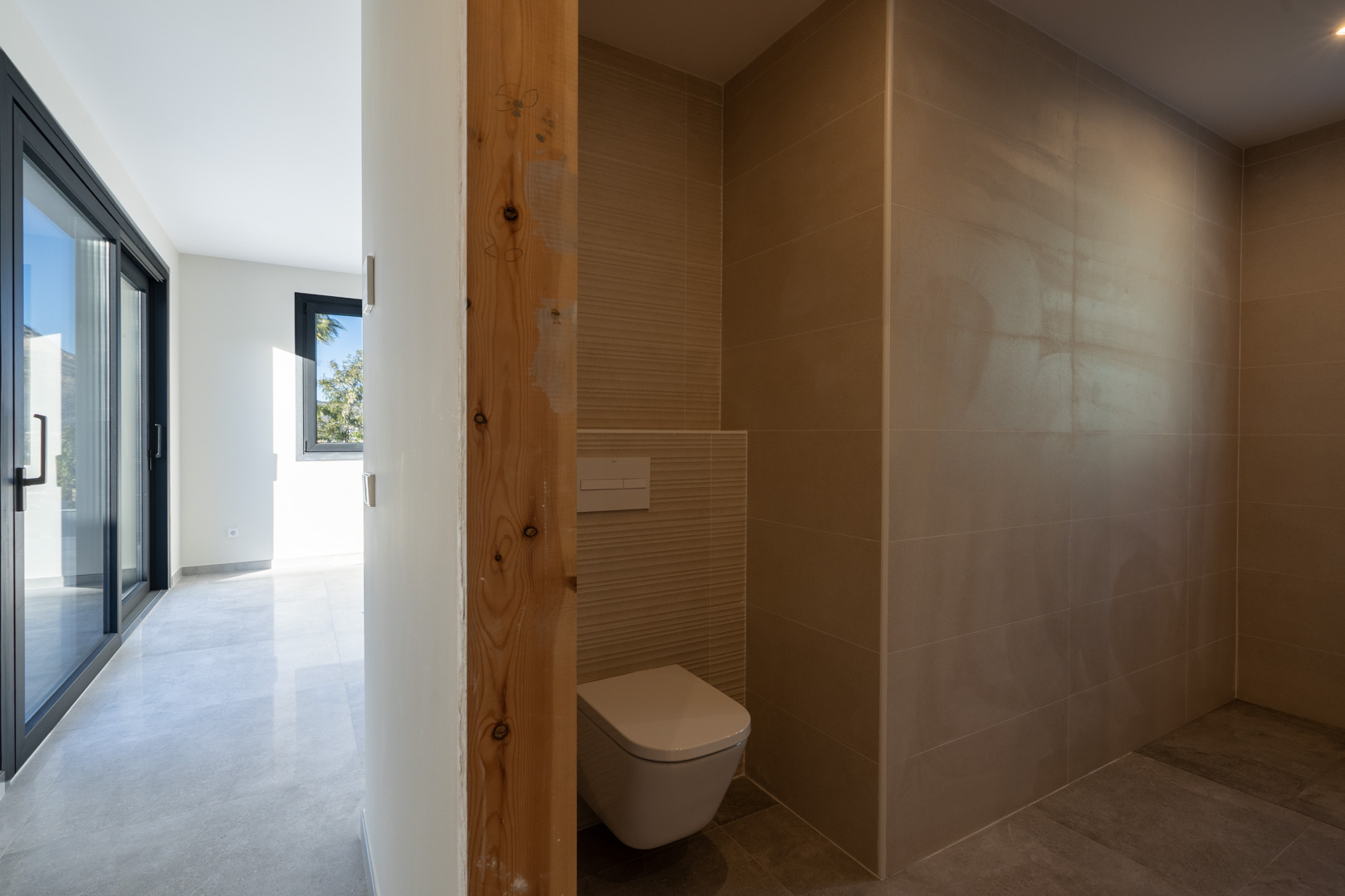 Détail des toilettes avec des carreaux couleur sable et un cadre de porte en bois brut, donnant sur le couloir.