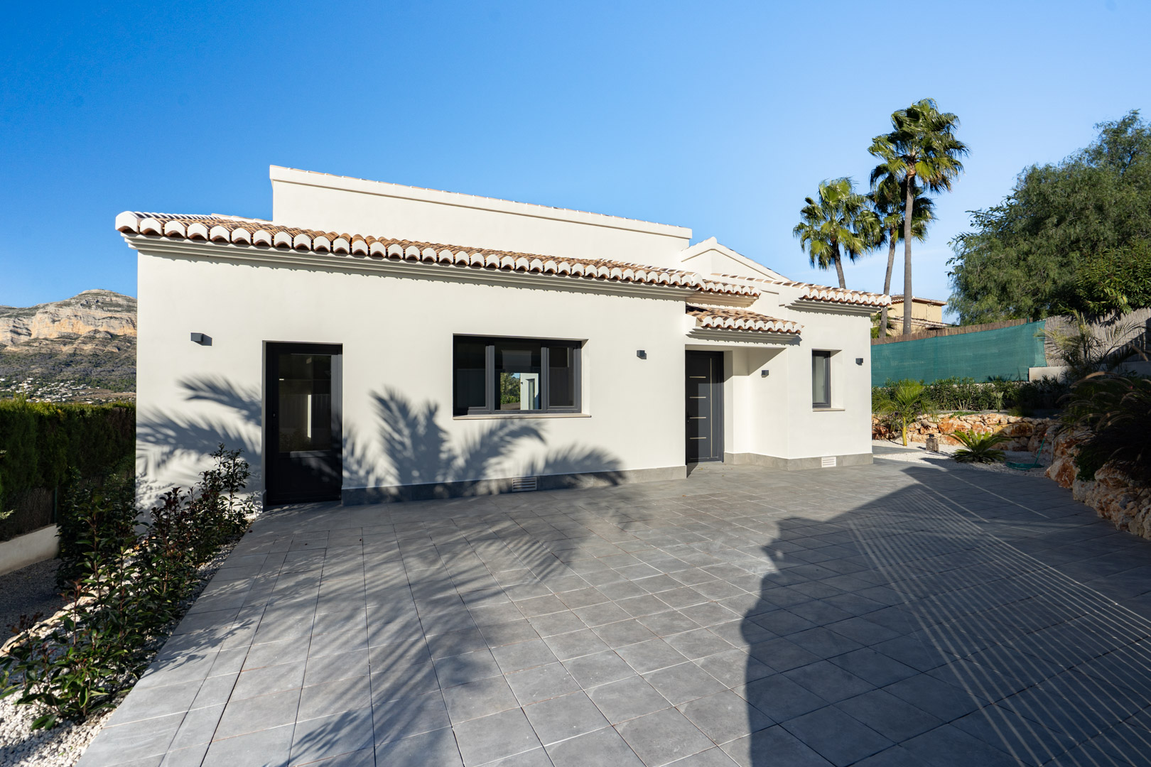 Extérieur de la villa avec façade blanche, tuiles en terre cuite et sol carrelé gris. Vue sur la montagne.