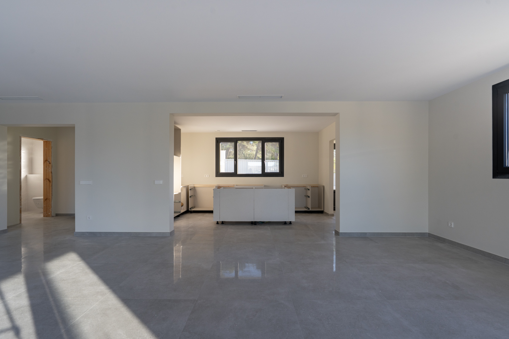 Intérieur décloisonné avec carrelage gris. Cuisine en construction avec meubles bas blancs et fenêtre à cadre noir.