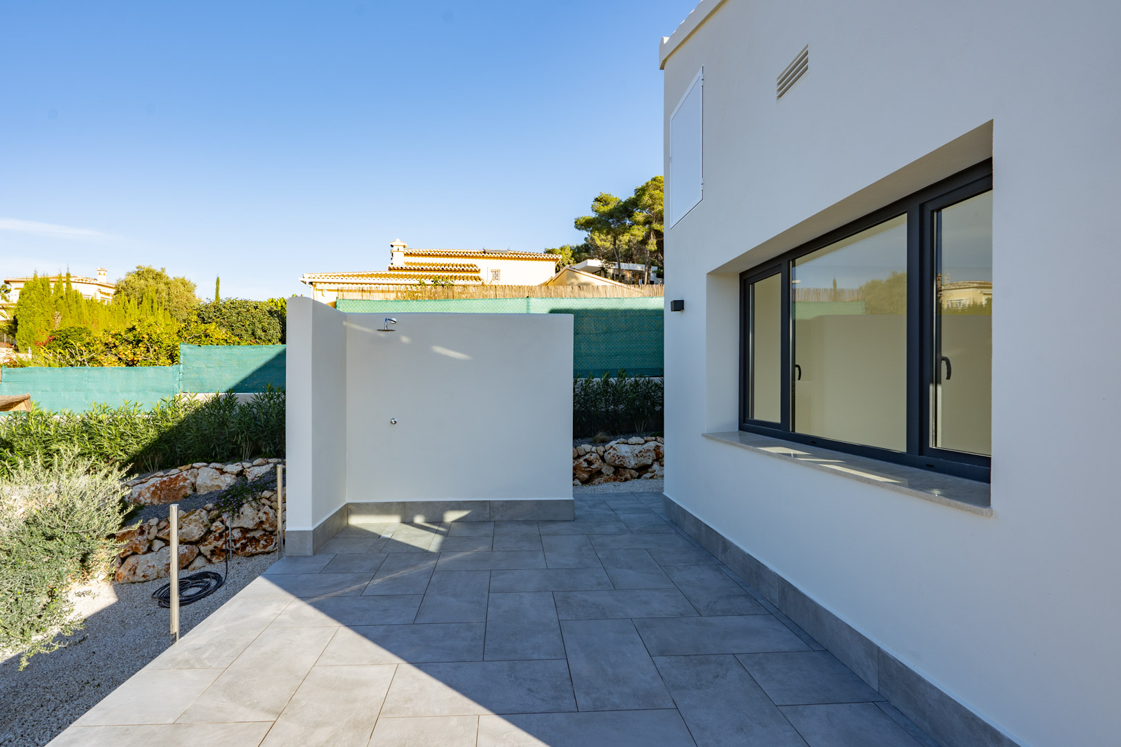 Patio avec douche extérieure et carrelage gris, à côté d'un mur blanc avec une fenêtre à double vitrage.