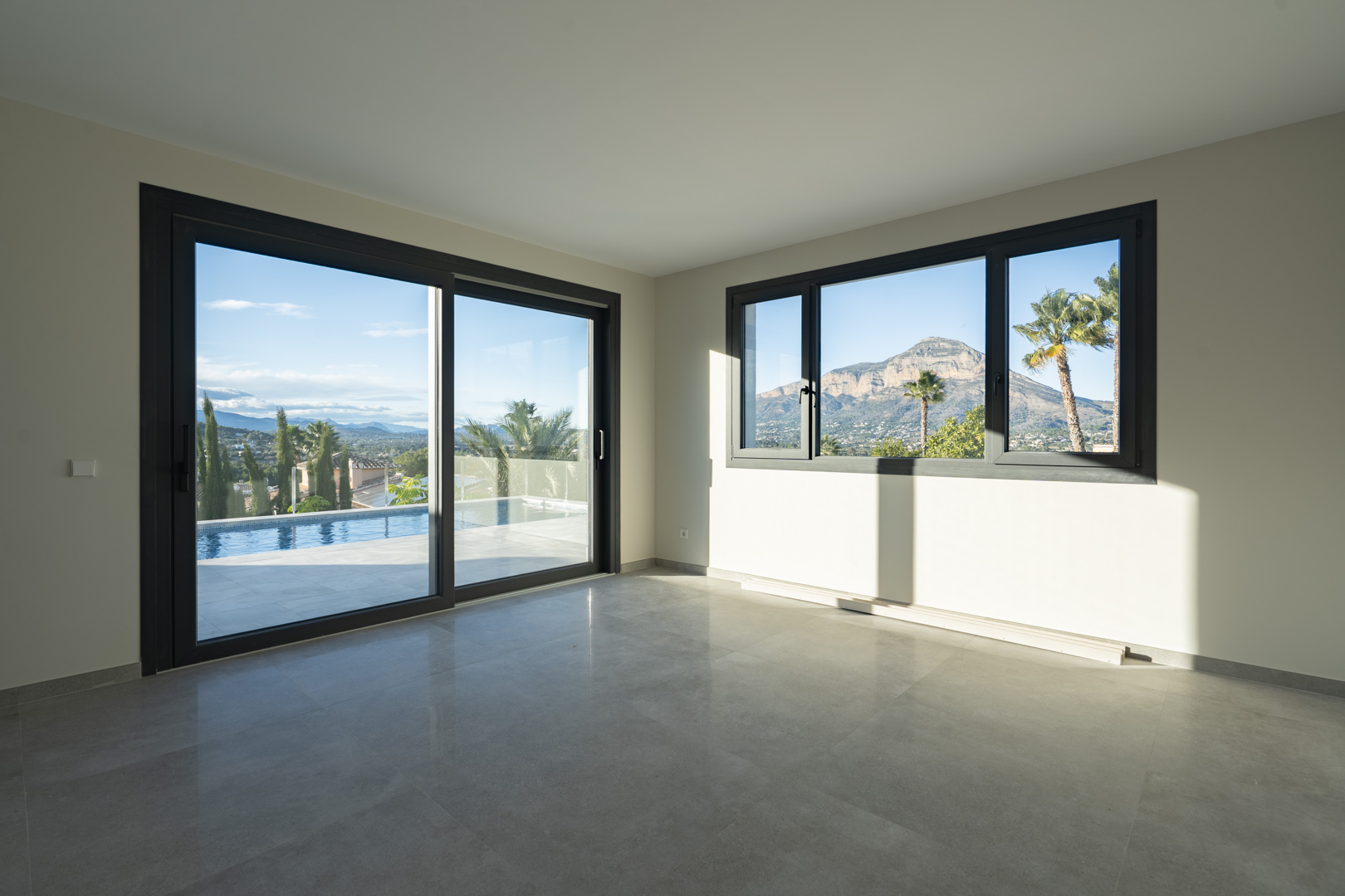 Salon spacieux avec carrelage gris et grandes fenêtres offrant une vue sur la piscine et le paysage montagneux.