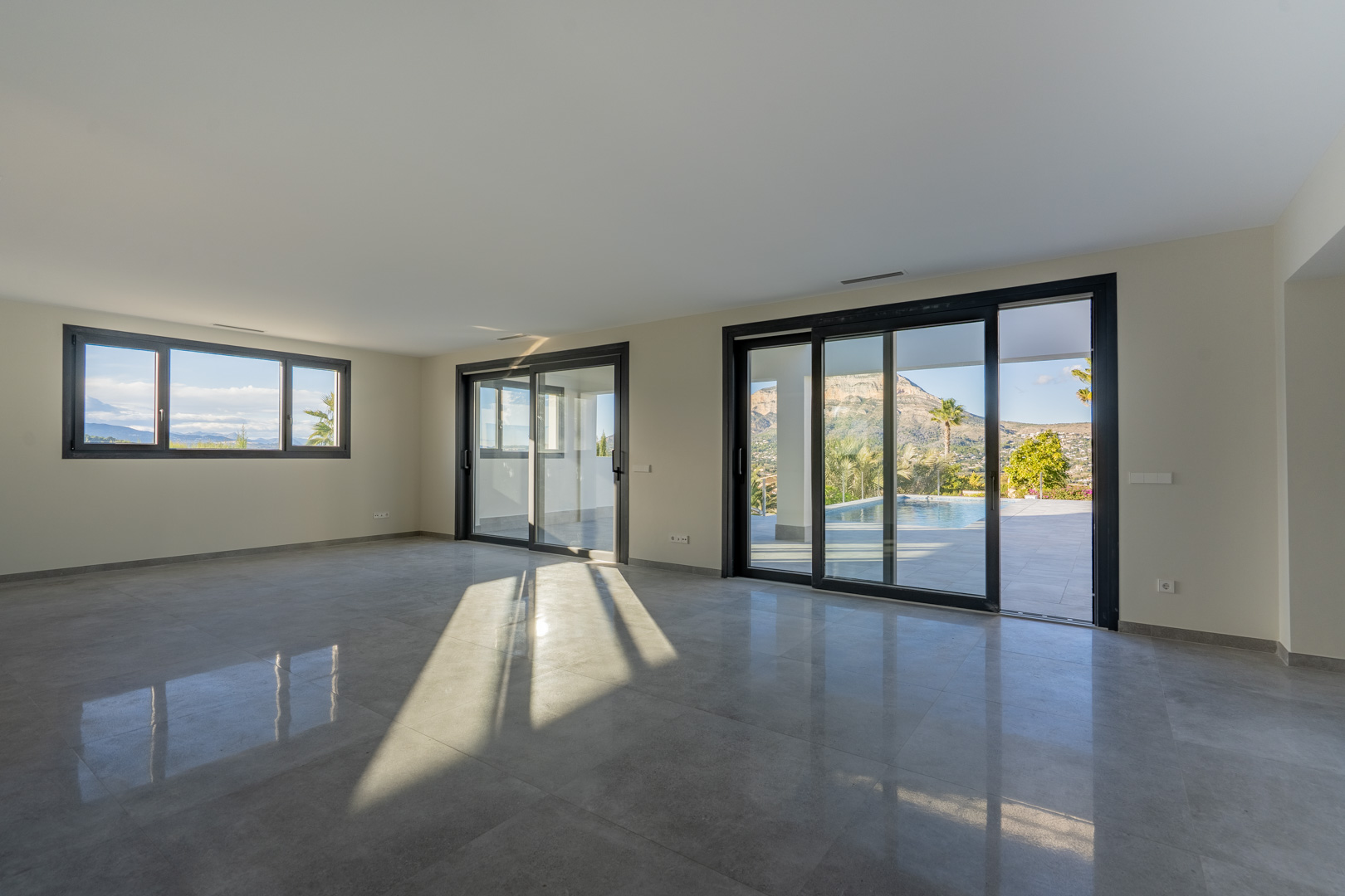 Salon spacieux avec carrelage gris et grandes fenêtres offrant une vue sur la piscine et le paysage.