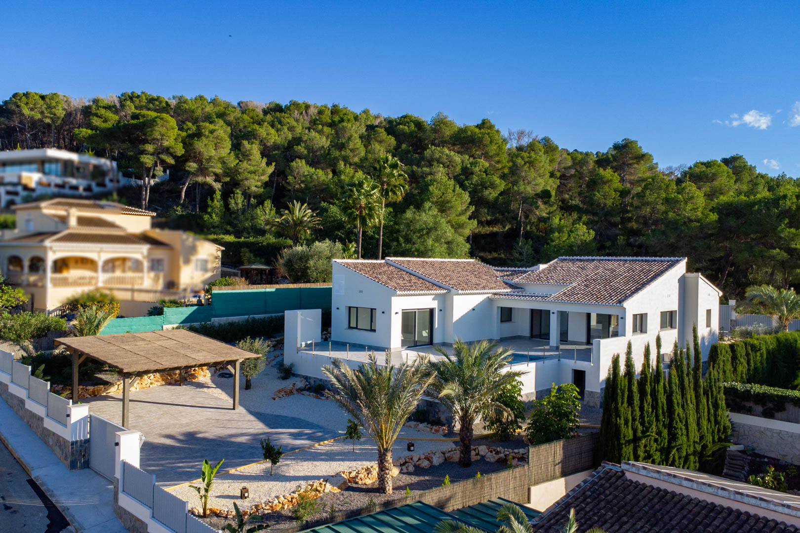 Vue aérienne d'une villa moderne avec des tuiles marron, une façade blanche et un abri voiture couvert à Jávea, entourée de verdure.