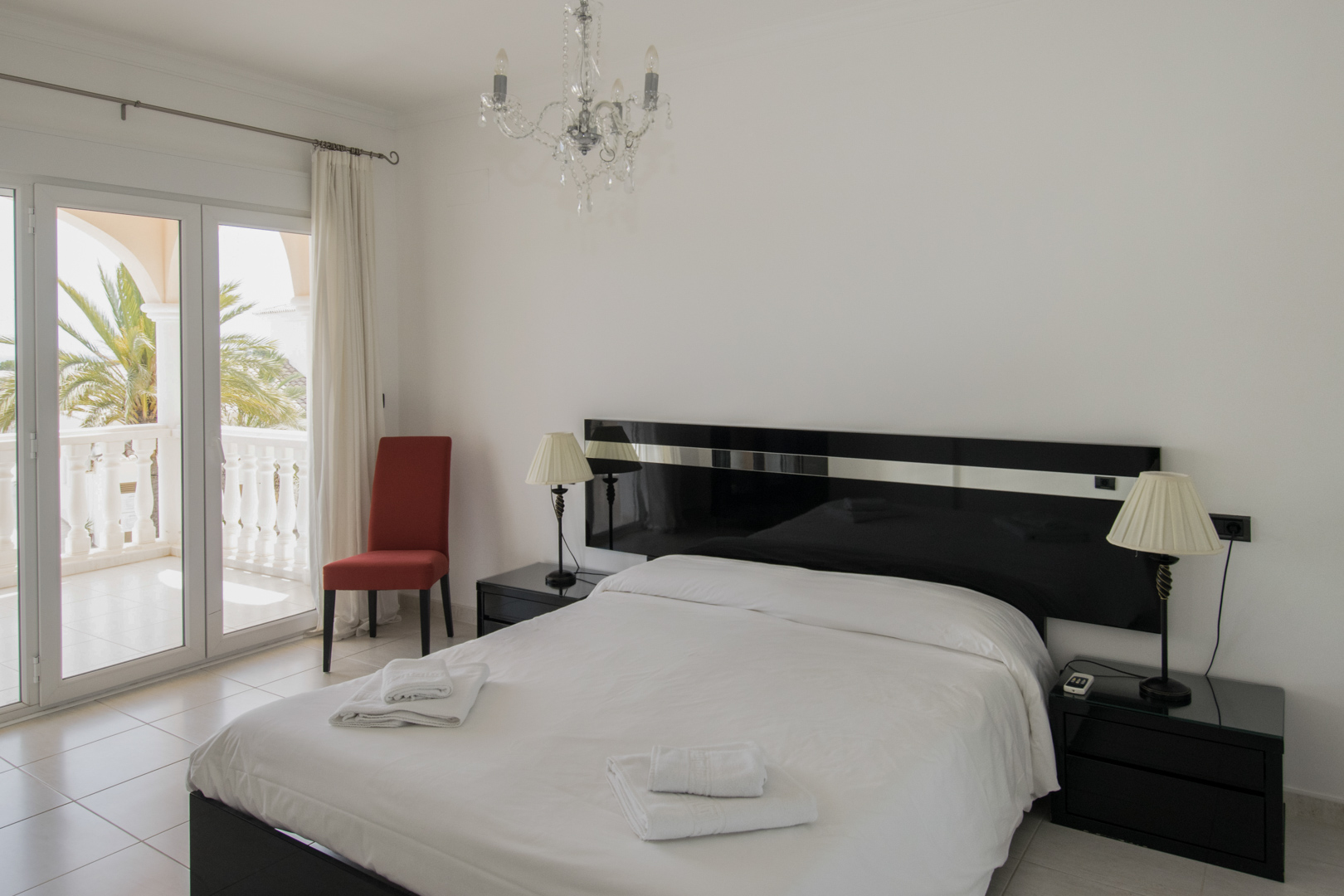 Chambre à coucher avec tête de lit laquée noire, lustre en cristal et accès direct à une terrasse privée avec vue sur les palmiers.