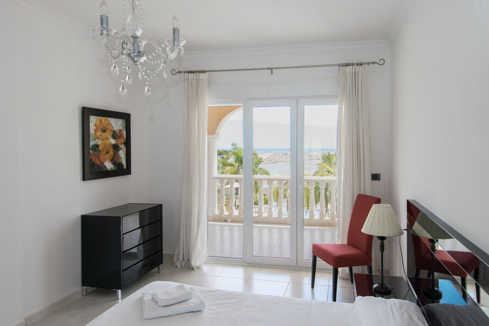 Chambre avec accès terrasse, commode noire moderne et vue mer. Elle dispose d'un lustre en cristal, d'un sol en carrelage clair et d'une chaise d'appoint rouge.