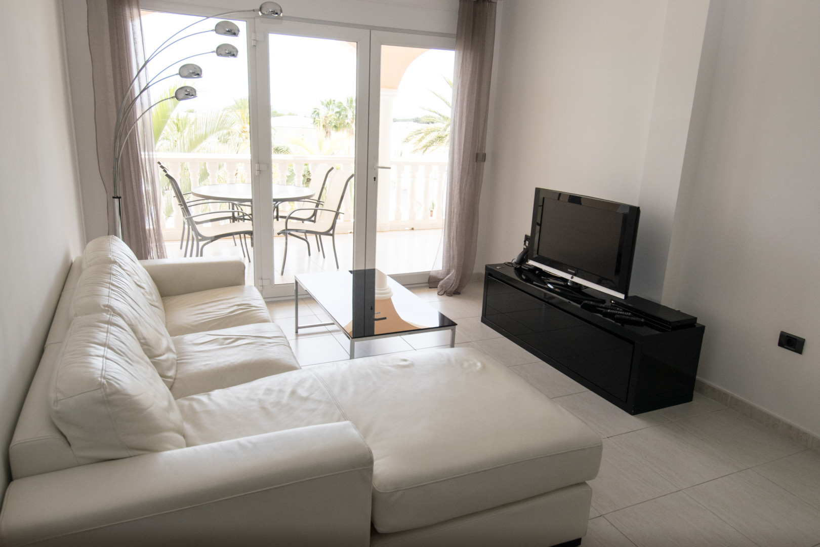 Espace de vie lumineux avec canapé en cuir blanc, meuble TV noir laqué et accès direct à une terrasse privée offrant une vue sur la végétation.