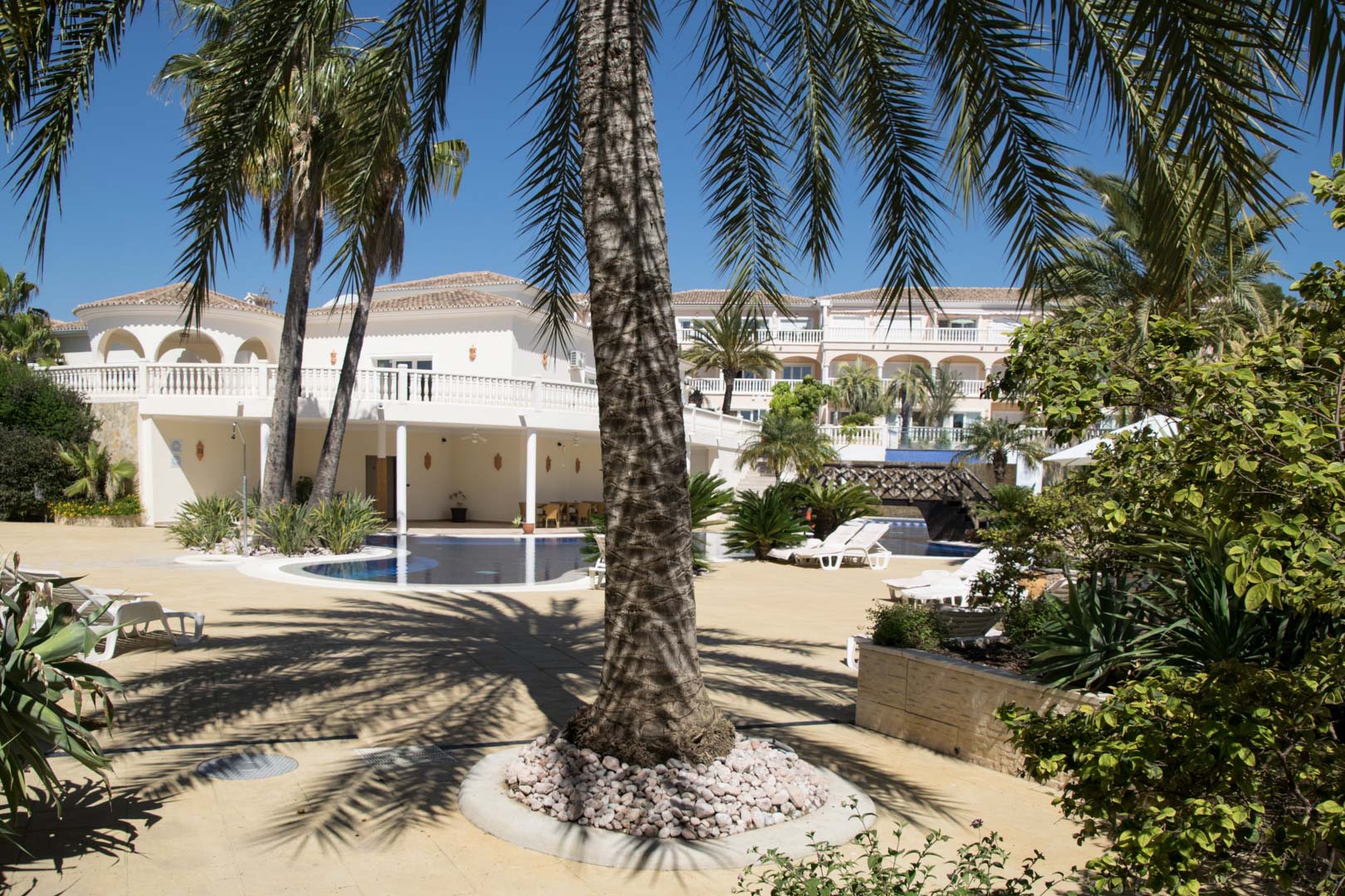 Espace extérieur avec piscine lagon, entouré de palmiers et de jardins méditerranéens. L'architecture propose des façades blanches avec arcades et terrasses.