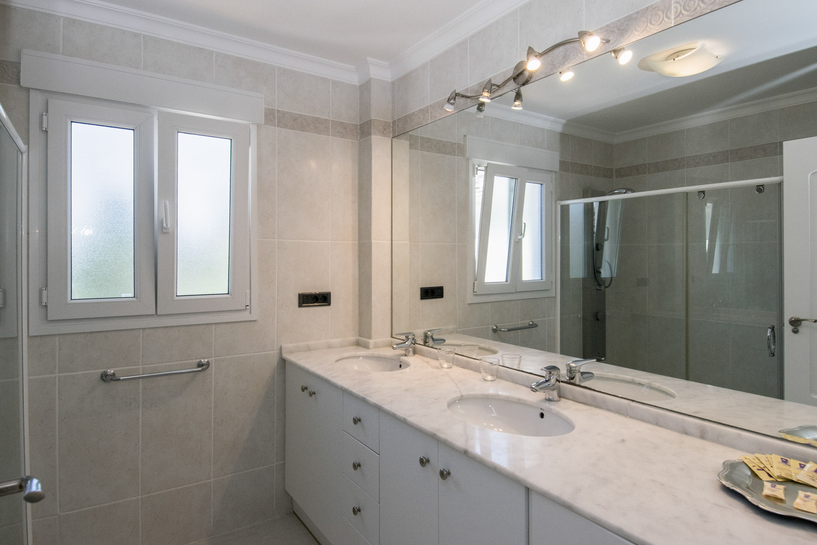 Salle de bain avec double vasque sur plan en marbre, meubles blancs et grand miroir éclairé. Murs carrelés dans des tons neutres et fenêtre oscillo-battante en verre dépoli.