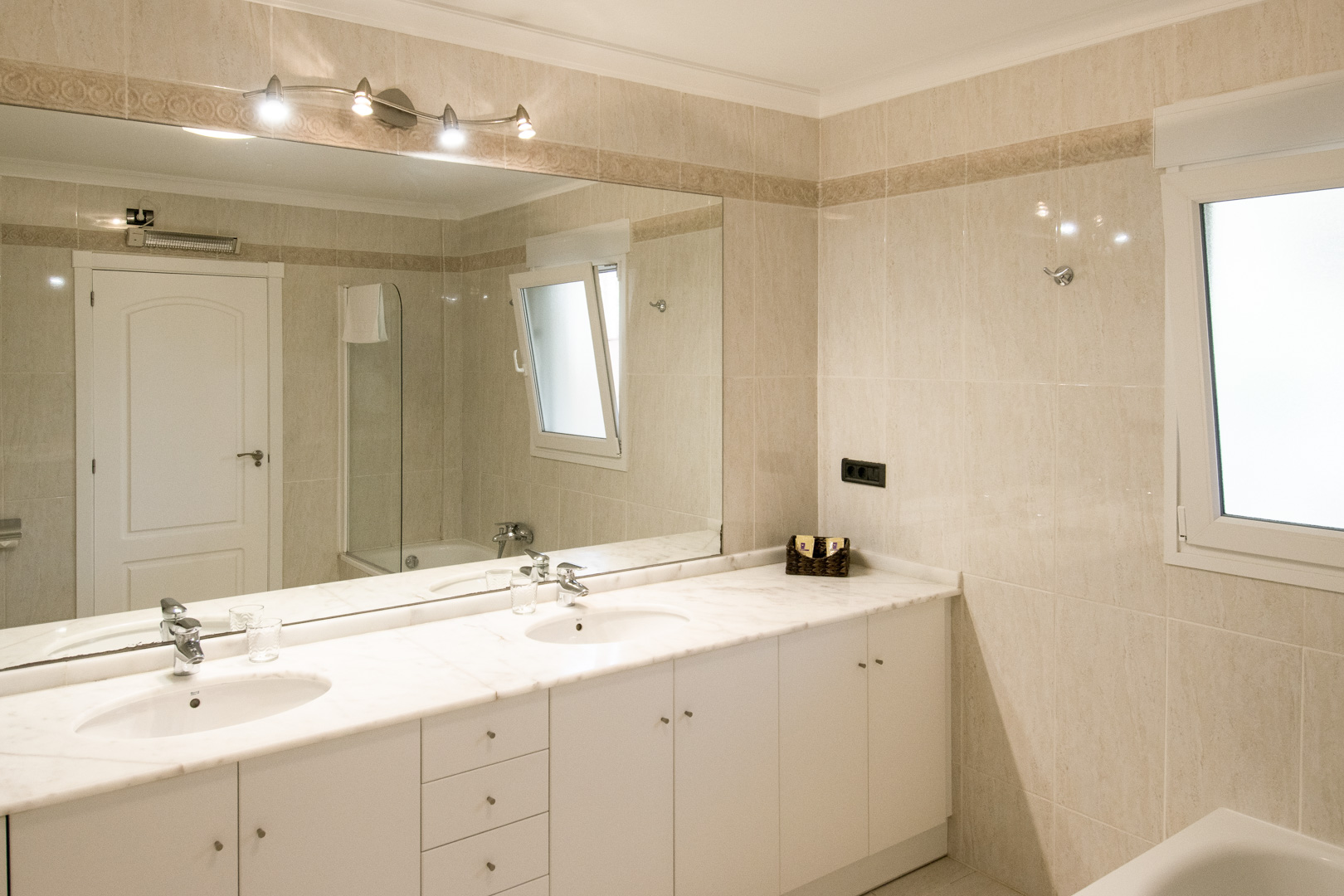 Salle de bain équipée d'un plan de travail en marbre blanc avec double vasque et grand miroir. Les murs sont revêtus de carrelage beige avec une frise décorative élégante.