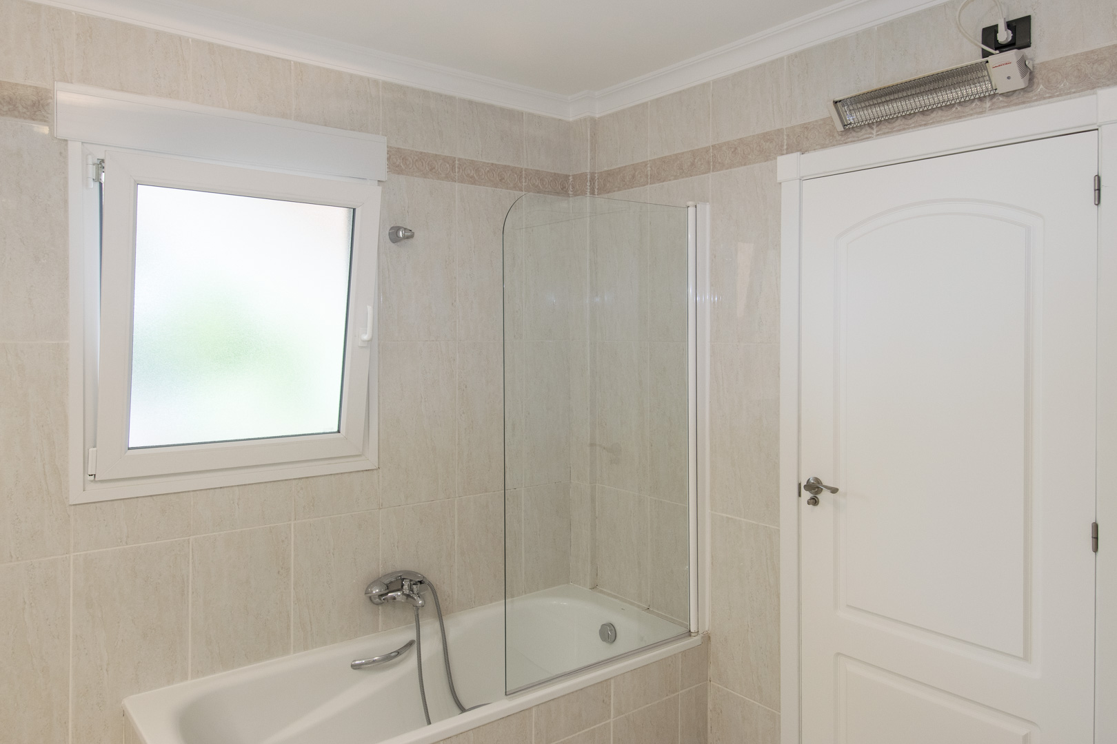 Salle de bains avec baignoire et paroi vitrée, murs carrelés avec frise décorative, fenêtre en PVC à verre dépoli et radiateur soufflant mural.