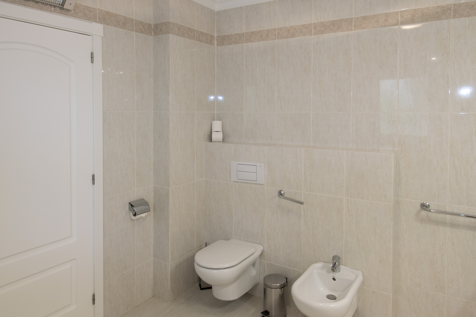 Salle de bains avec carrelage beige et frise décorative, équipée d'un WC suspendu, d'un bidet et de barres d'appui chromées.