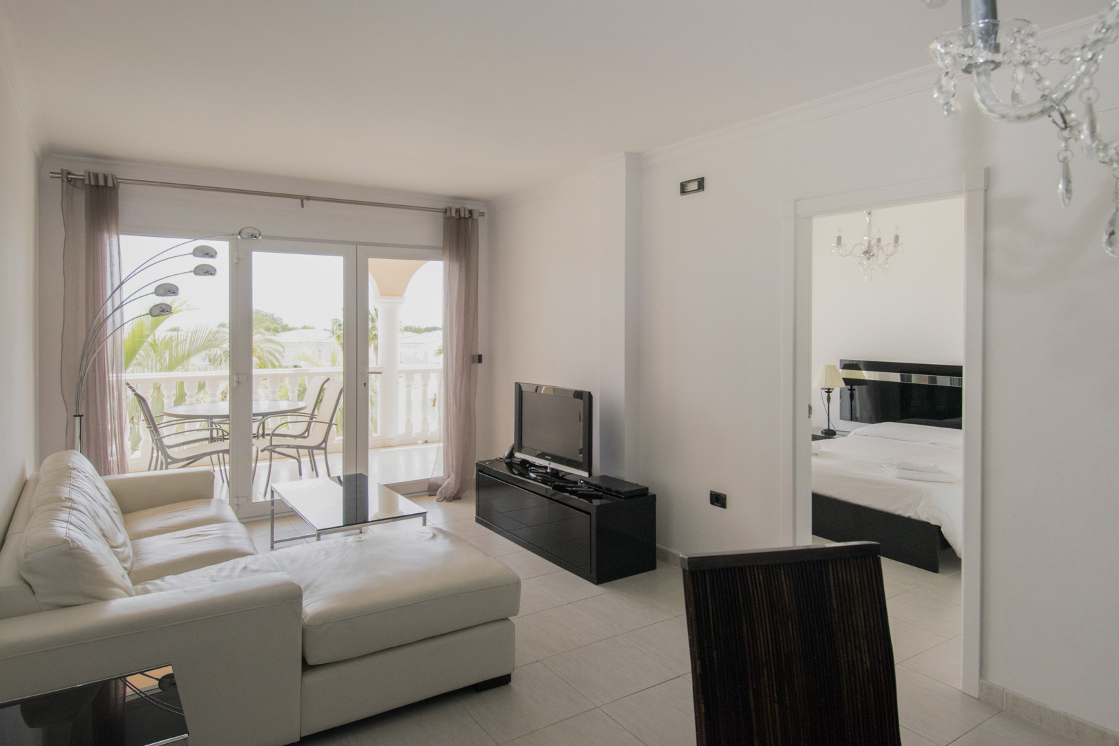 Salon avec canapé en cuir blanc, mobilier noir et accès terrasse. L'espace se prolonge vers la chambre, souligné par des lustres en cristal et un sol en carrelage clair.