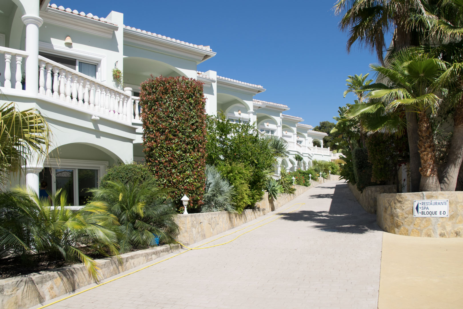 Vue extérieure du complexe méditerranéen avec balustrades blanches, terrasses voûtées et jardins luxuriants bordant une allée pavée sous un ciel dégagé.