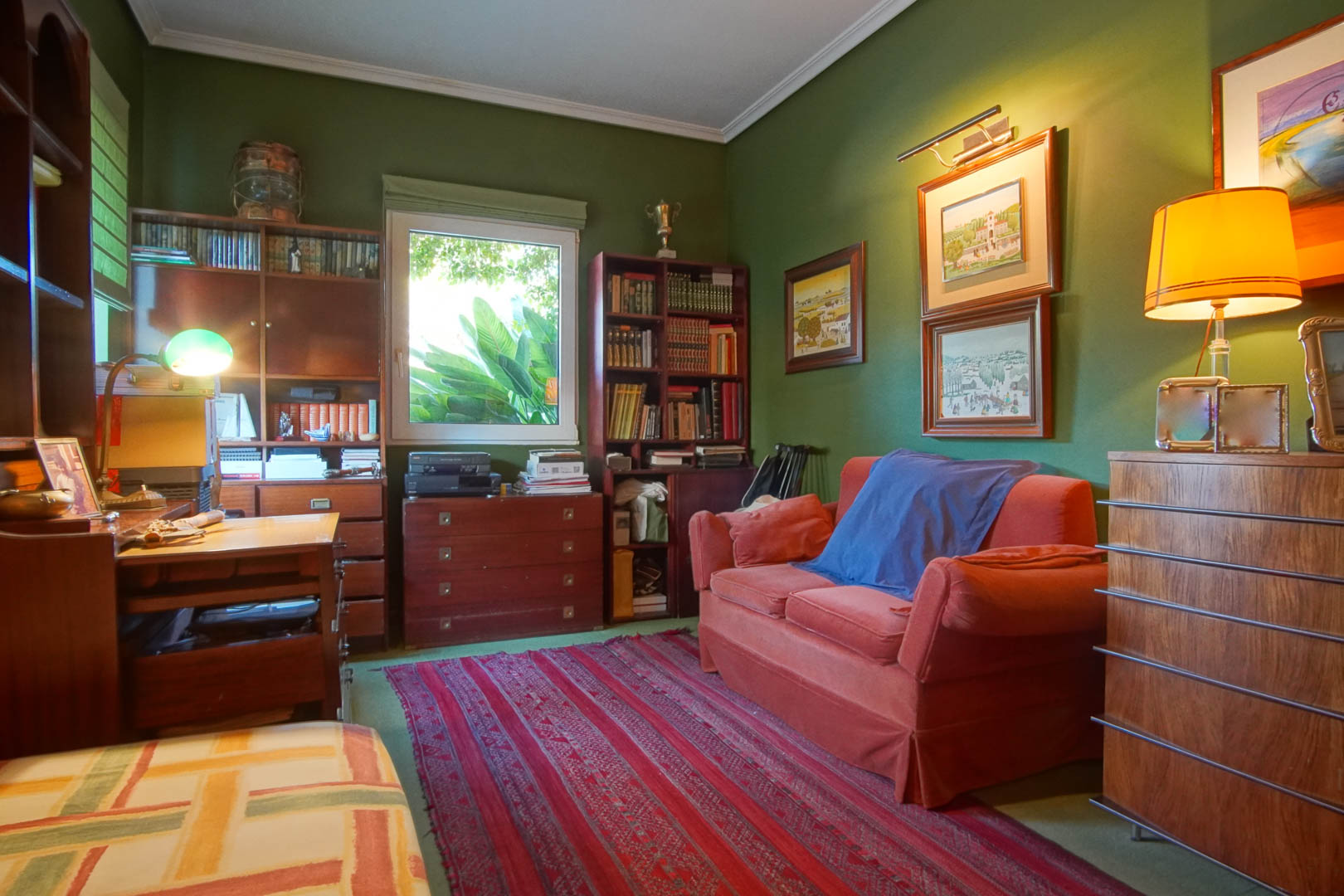 Bureau aux murs verts et mobilier en bois classique. Il dispose d'étagères, d'un bureau, d'un canapé rouge et d'une lumière naturelle avec vue sur la verdure.