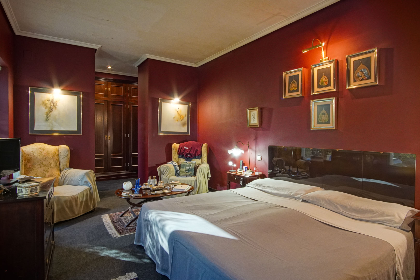 Chambre aux murs bordeaux avec coin salon, fauteuils et armoires encastrées en bois sombre. L'éclairage ciblé souligne les œuvres d'art et l'élégance de la pièce.