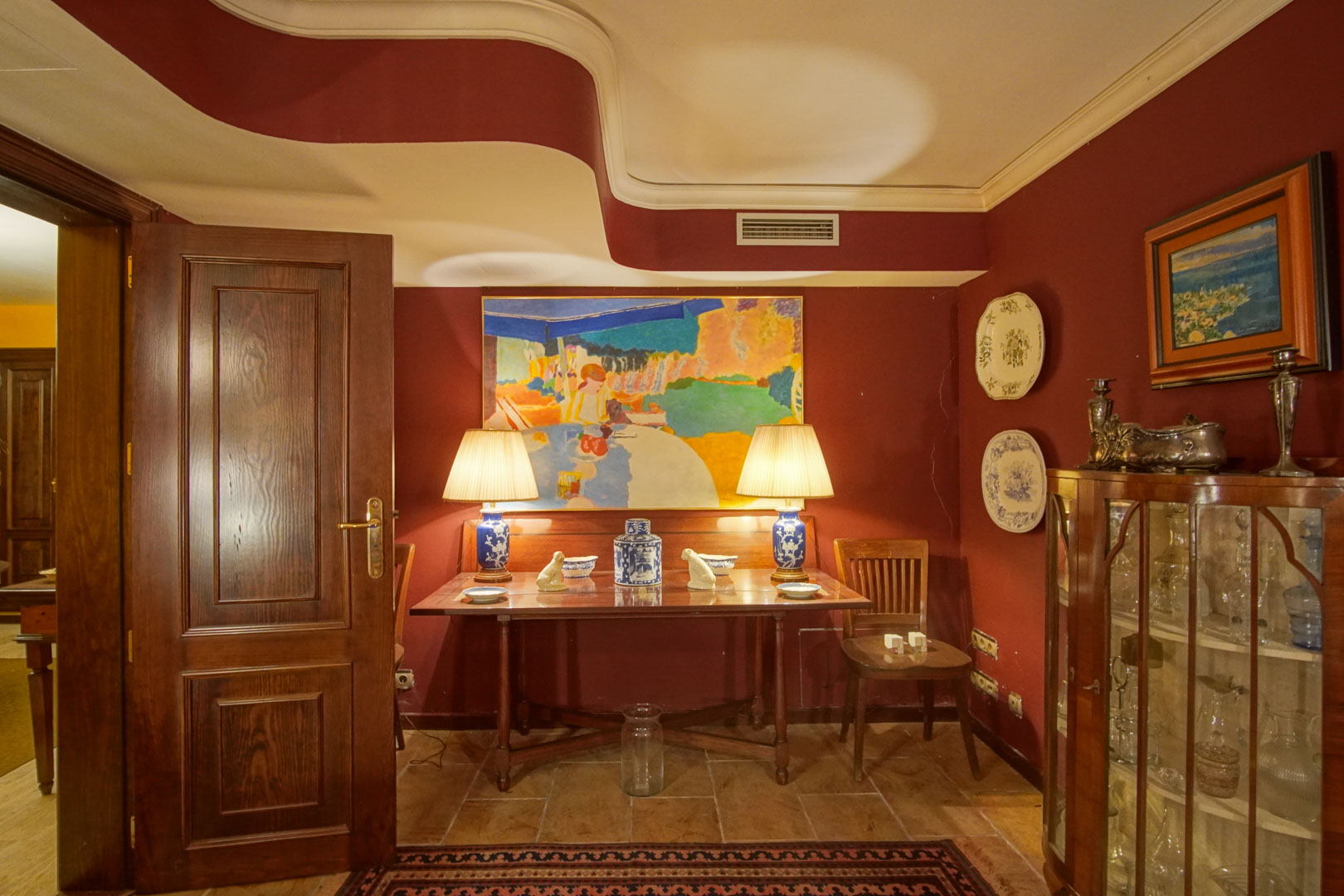 Coin élégant avec murs bordeaux, plafond aux lignes ondulées et mobilier en bois classique. Comprend une vitrine d'exposition et des lampes en céramique sur un sol en pierre naturelle.
