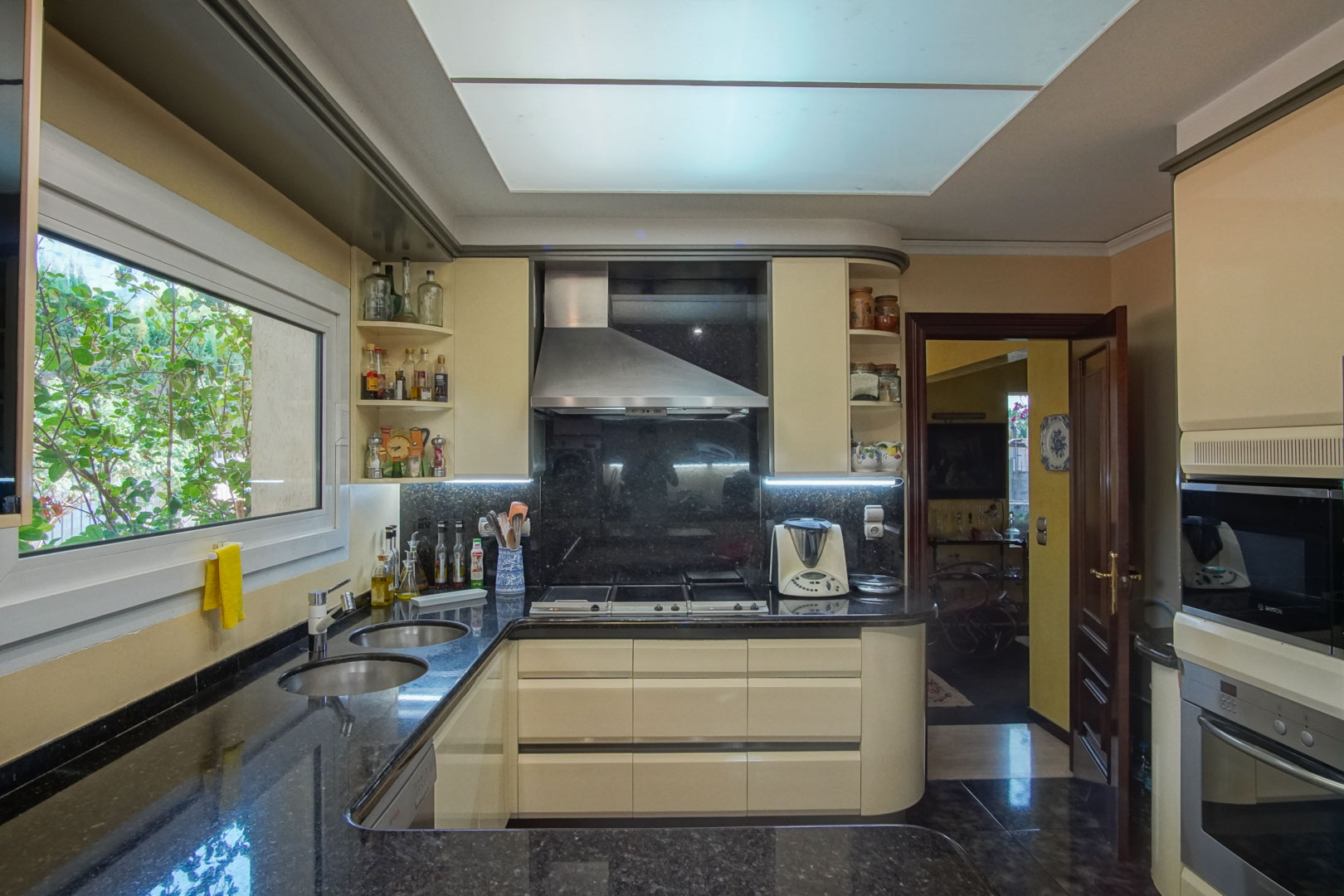 Cuisine indépendante avec plans de travail en granit noir, meubles crème et électroménagers intégrés, bénéficiant d'une grande fenêtre et d'un éclairage zénithal.
