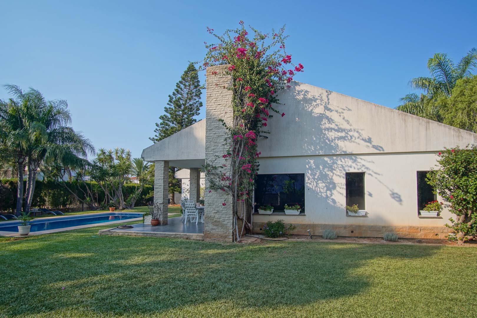Façade extérieure avec cheminée en pierre et bougainvillier, jardin méditerranéen avec pelouse naturelle et piscine privée sous un ciel azur.