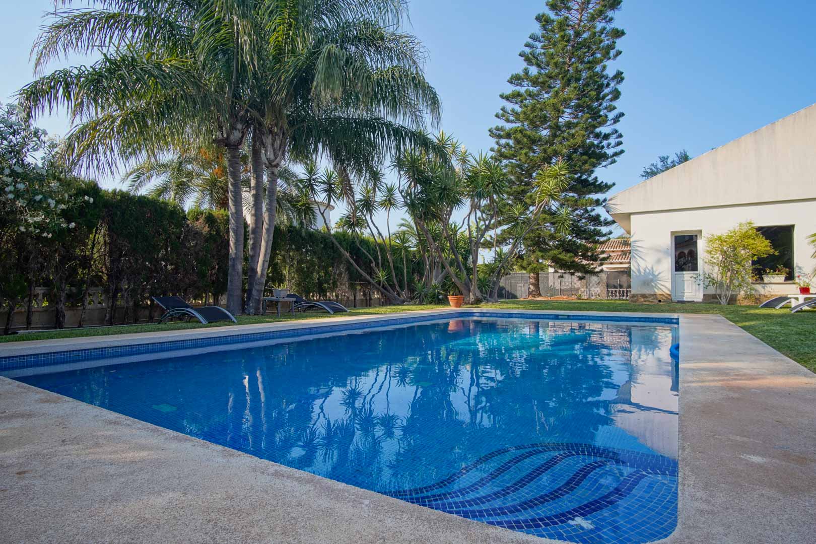 Piscine privée avec revêtement en mosaïque bleue, entourée d'un jardin méditerranéen avec palmiers, pelouse naturelle et une haie assurant une intimité totale.