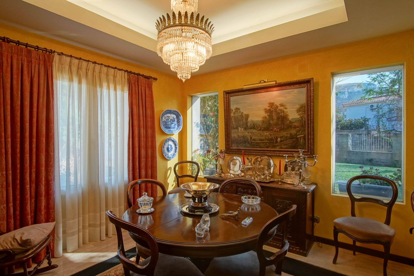 Salle à manger élégante avec table ronde en bois, lustre en cristal et buffet classique. Les murs ocre, le plafond à caissons et les vues sur le jardin complètent cet espace raffiné.