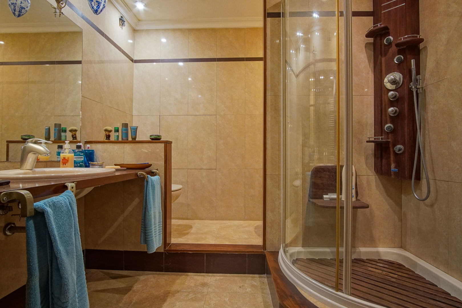 Salle de bain avec carrelage en marbre beige, plan de travail en bois et cabine de douche courbe équipée d'une colonne d'hydromassage.