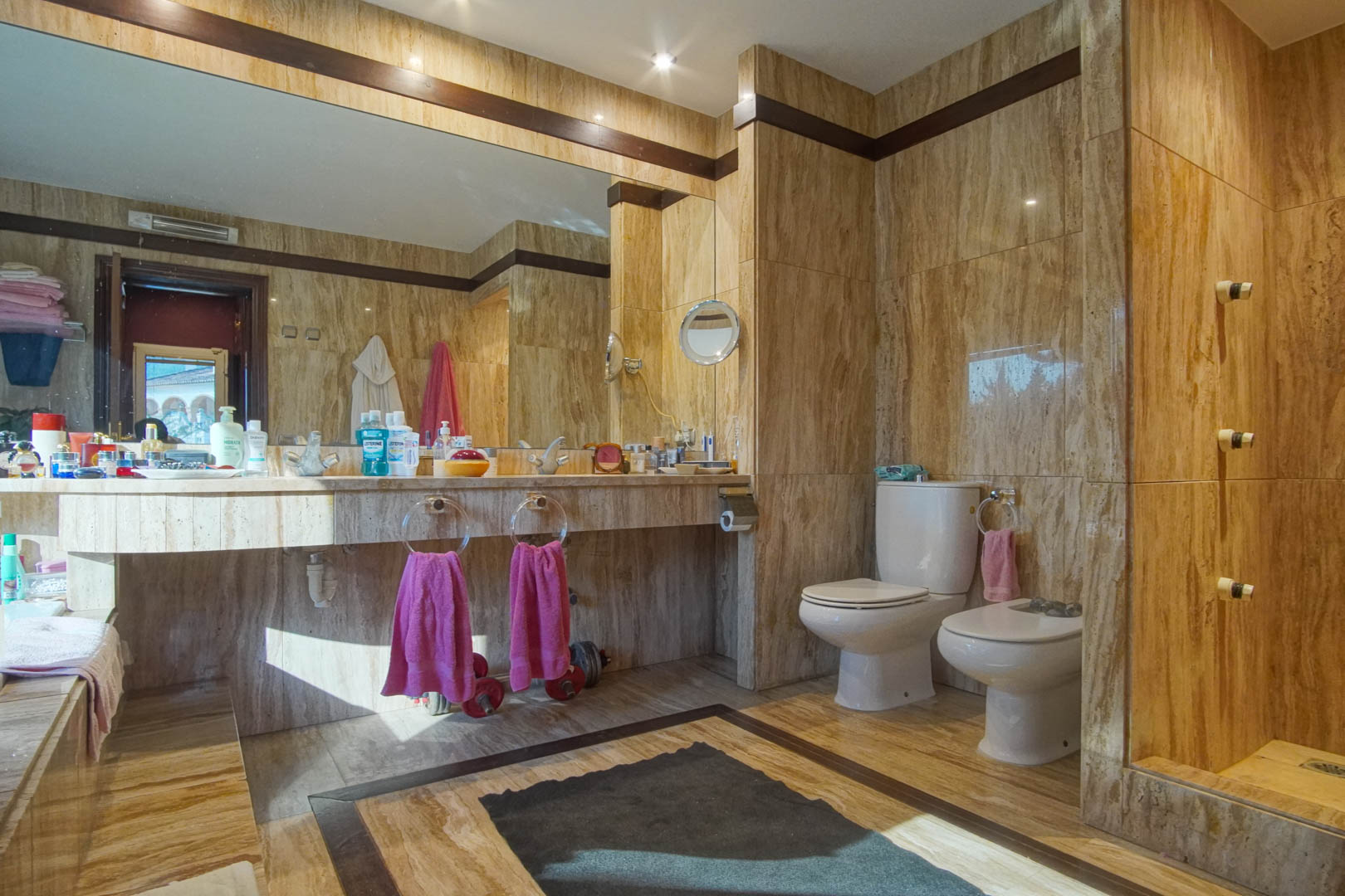 Salle de bain luxueuse en marbre travertin avec vasque intégrée, grand miroir mural, éclairage encastré et finitions élégantes en bois sombre.