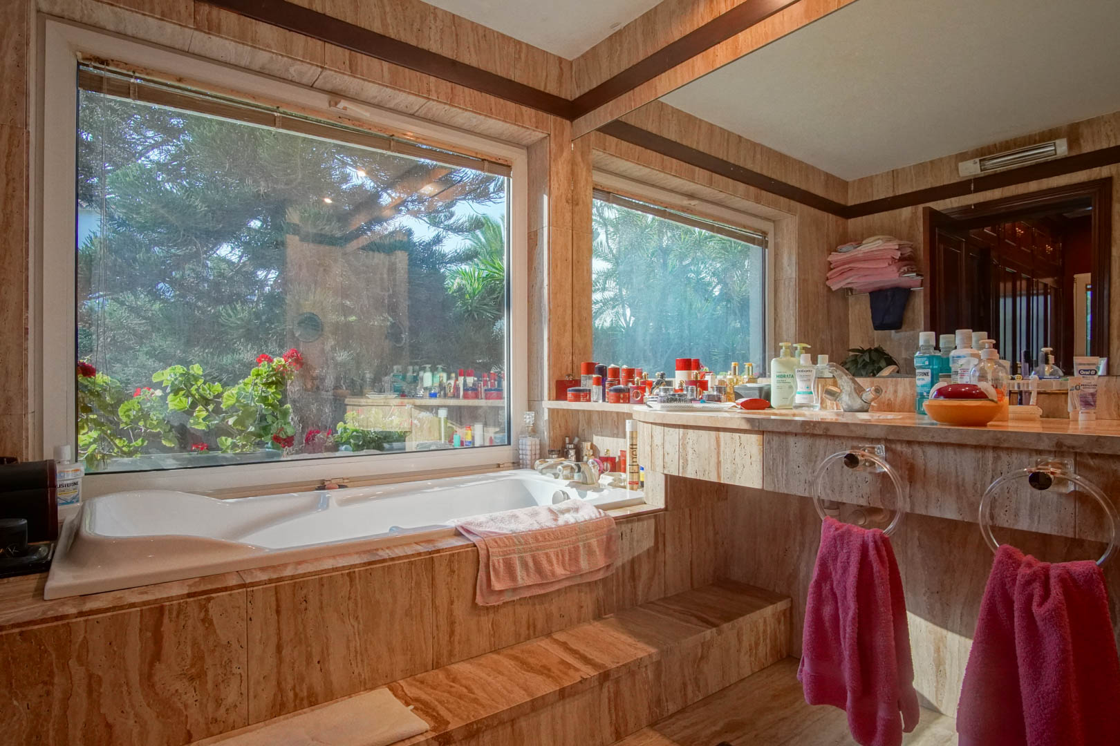 Salle de bain revêtue de marbre travertin avec baignoire encastrée, grande fenêtre donnant sur le jardin et plan de toilette intégré avec grand miroir.