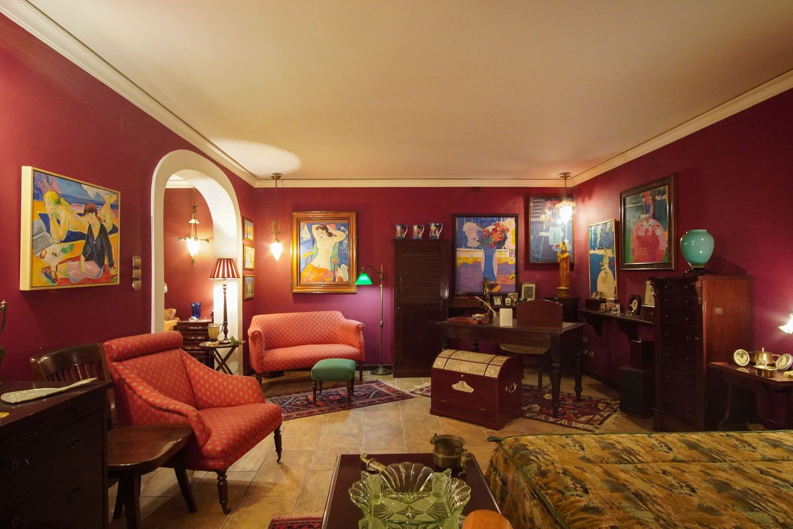 Salon classique aux murs bordeaux et sols en marbre, comprenant un coin bureau. La pièce se distingue par sa collection d'art, son mobilier d'époque et son éclairage chaleureux.