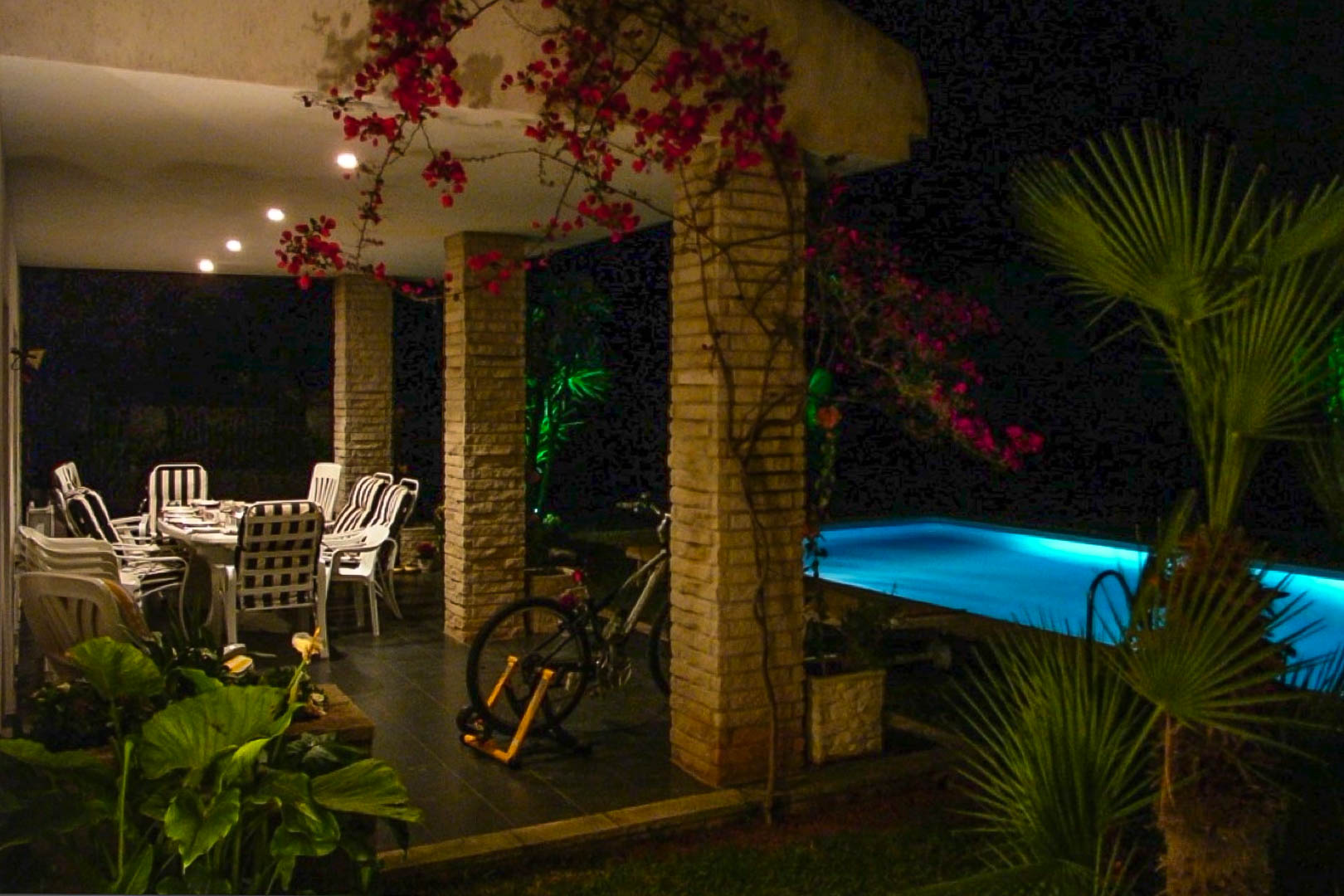 Terrasse couverte avec colonnes en briques apparentes, coin repas extérieur et bougainvilliers, bordant une piscine éclairée en soirée.